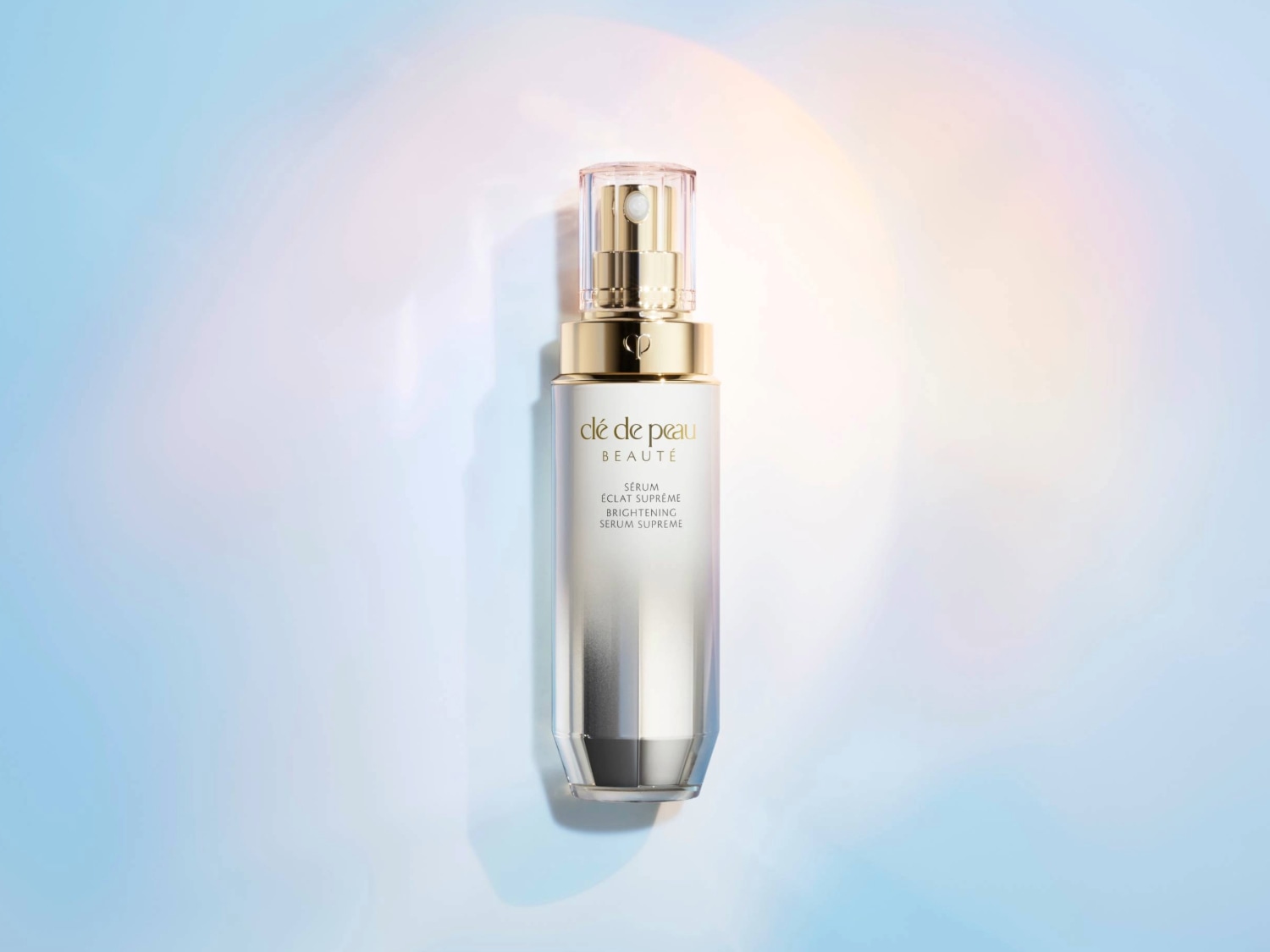 Voici le sérum éclat suprême de Clé de Peau Beauté dans son flacon dégradé argenté et doré sur un fond bleu et rose pastel.