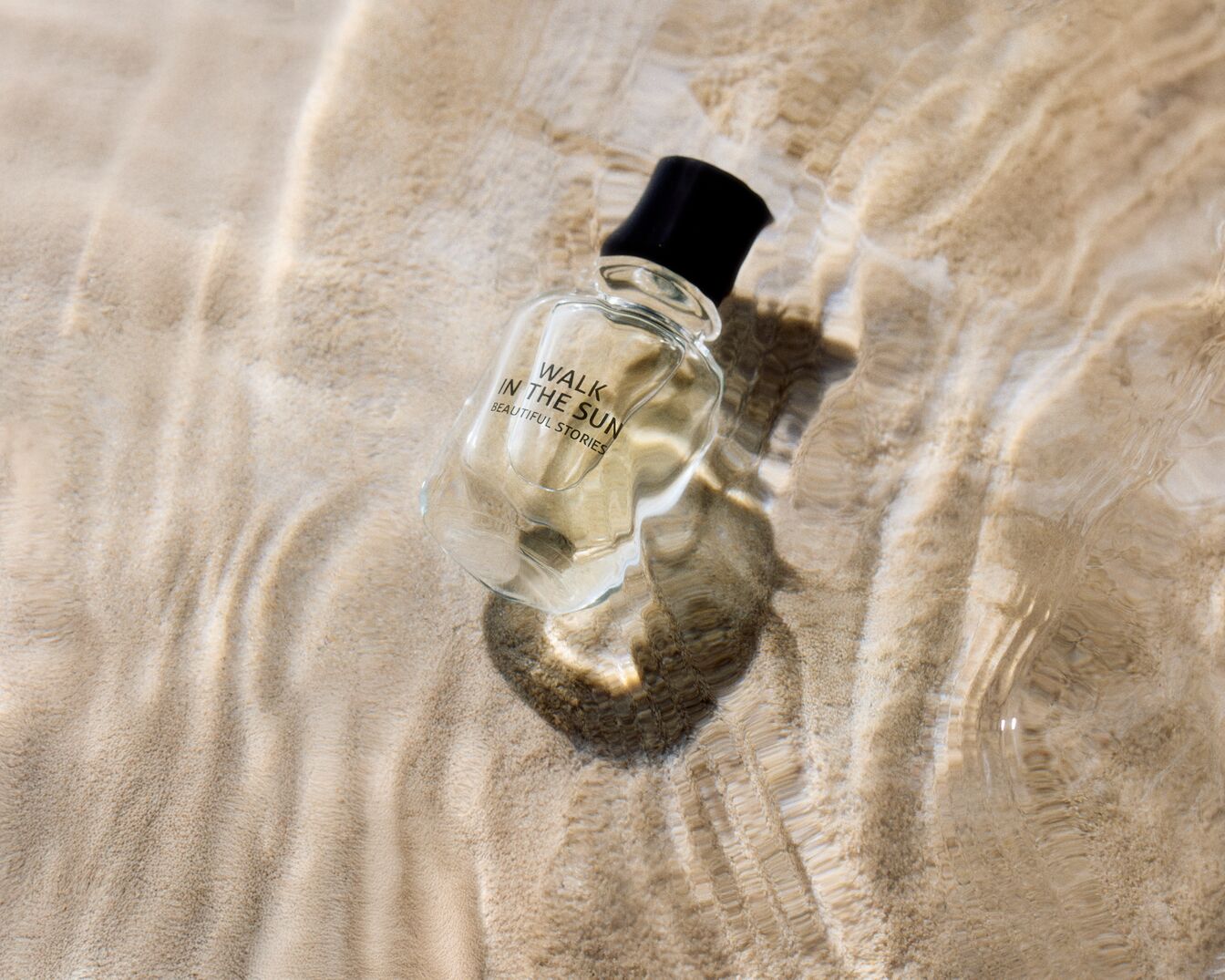Un flacon transparent de parfum "Walk in the Sun Beautiful Stories" repose sur du sable beige clair, partiellement immergé dans de l'eau claire.