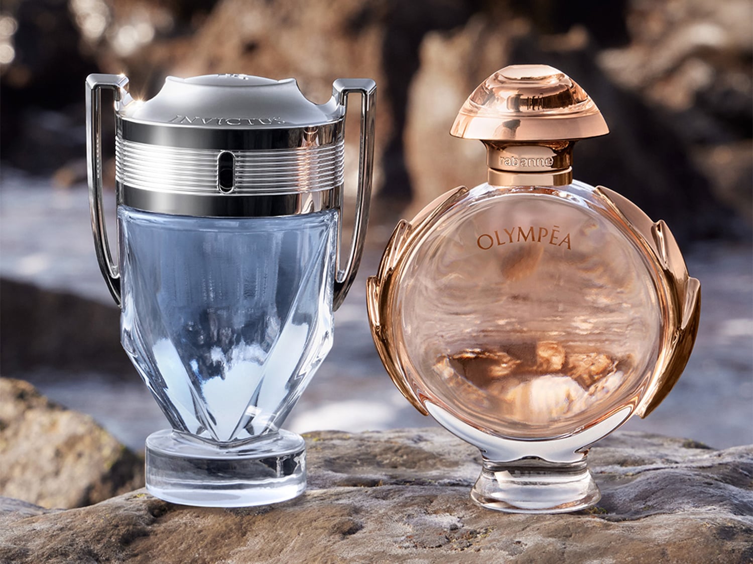 Zwei Parfümflaschen von Paco Rabanne stehen auf einem Felsen: Invictus, ein blauer Duft in einem silbernen Pokalflakon, und Olympéa, ein roségoldener Duft in einem runden Flakon mit goldenen Flügeln.