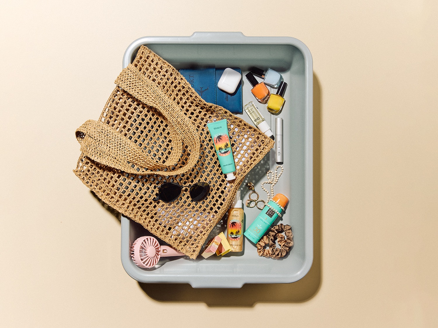 Vue aérienne d'un sac de plage en osier rempli de produits de beauté, de lunettes de soleil et d'un ventilateur rose dans un bac de sécurité gris.