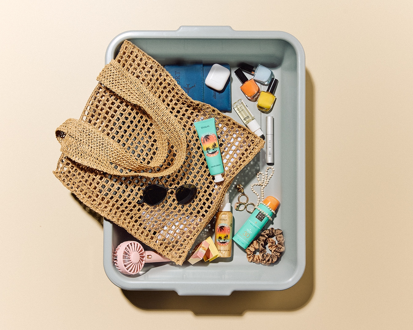 Dans un plateau gris, un sac de plage en paille tressée côtoie des essentiels de beauté : crème turquoise, solaires, vernis colorés, lunettes de soleil et un mini ventilateur rose.