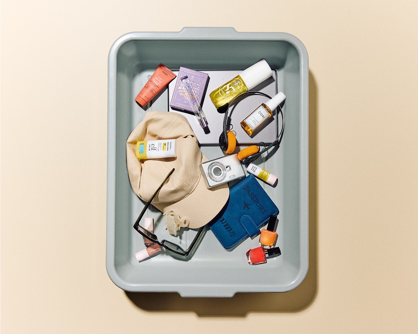 Dans un bac de sécurité aéroportuaire, on retrouve un passeport bleu, un chapeau beige, des écouteurs noirs et plusieurs produits de beauté.