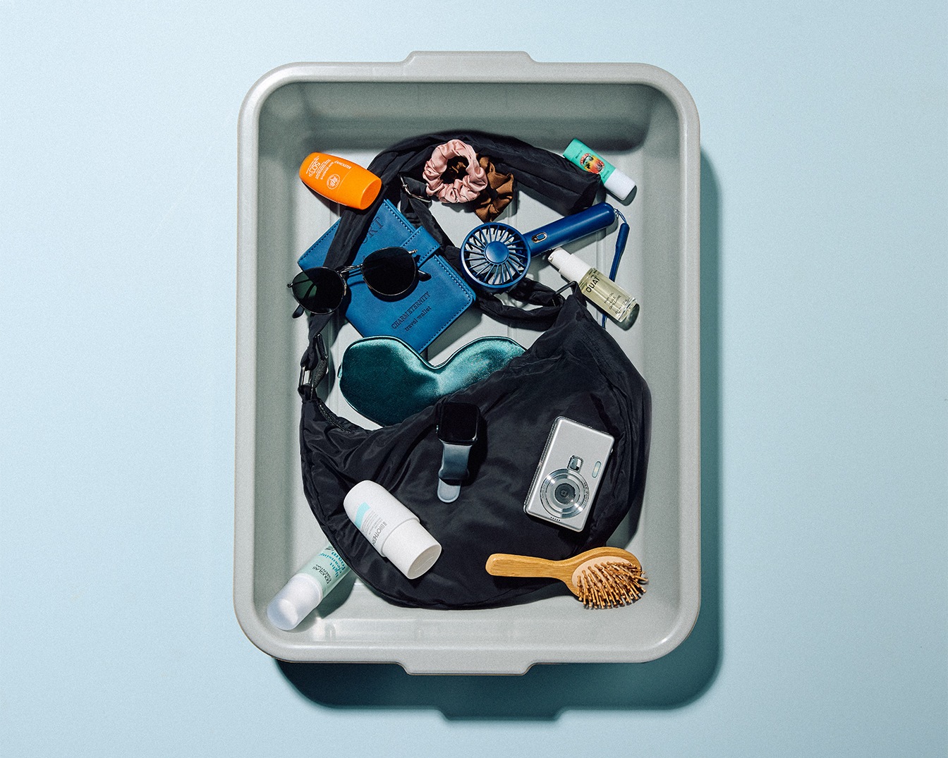 Dans un bac de sécurité aéroportuaire, on retrouve un sac noir rempli d'essentiels de voyage tels que lunettes de soleil, crème solaire orange, portefeuille bleu, et une mini-caméra argentée.