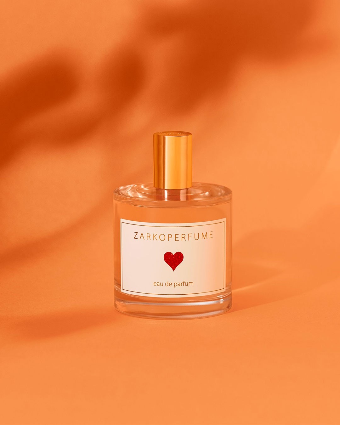 Présentation d'un flacon de parfum ZARKOPERFUME, avec un cœur rouge sur l'étiquette, sur un fond orange vif, évoquant une fragrance à la fois douce et passionnée.