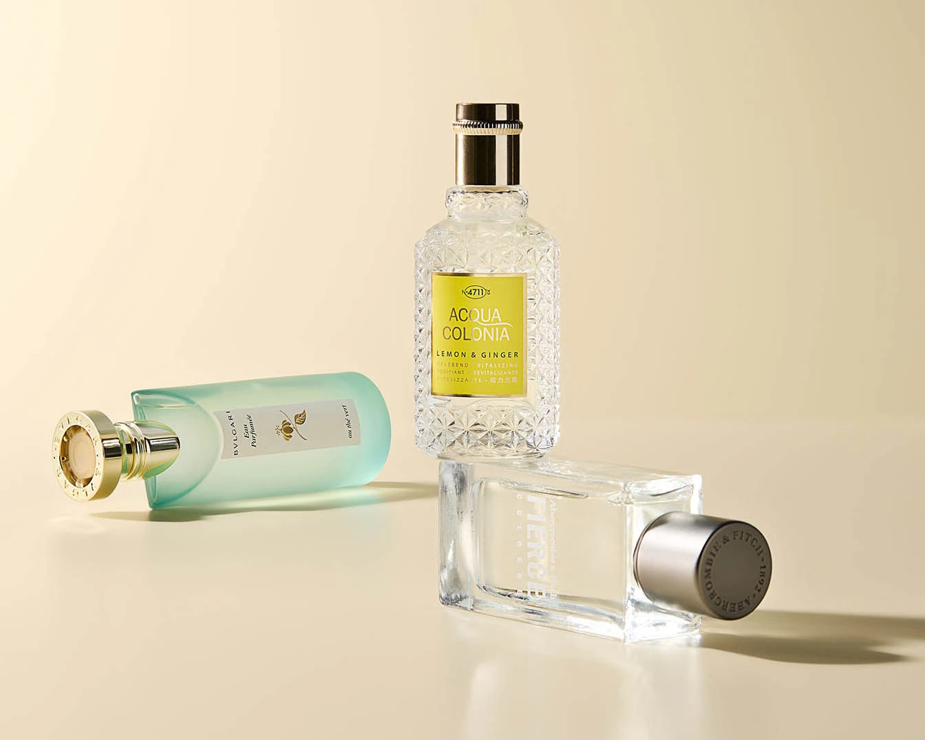 Drie flessen parfum staan op een beige achtergrond, waaronder Bvlgari Eau Parfumée au thé vert, 4711 Acqua Colonia Lemon & Ginger en Abercrombie & Fitch Fierce.