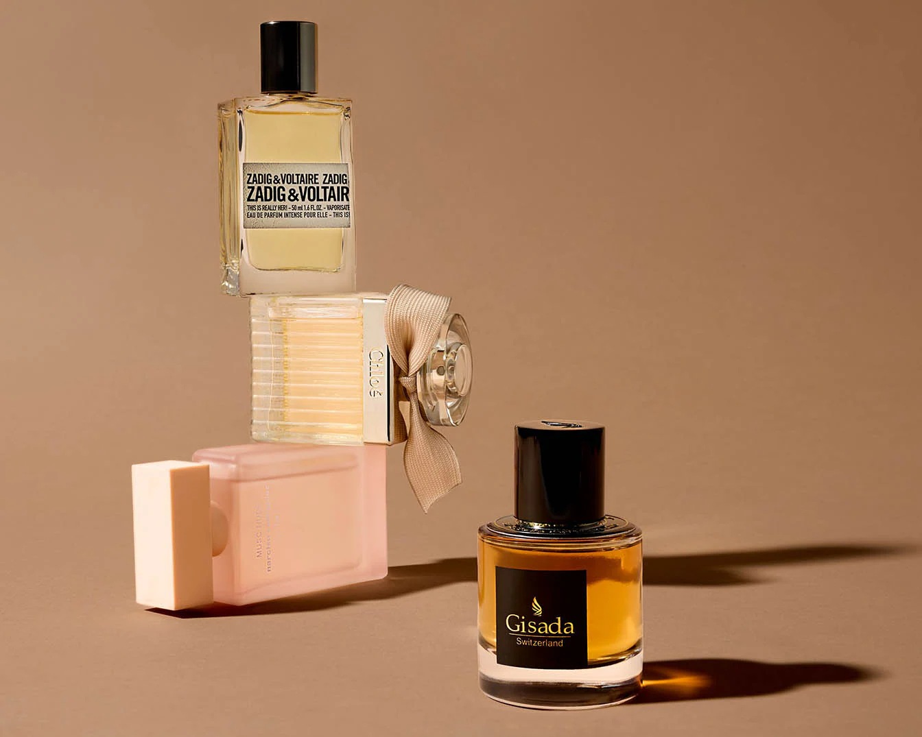 Vier parfumflessen van verschillende merken, waaronder Zadig&Voltaire, Chloé en Gisada, gestapeld op een bruine achtergrond.