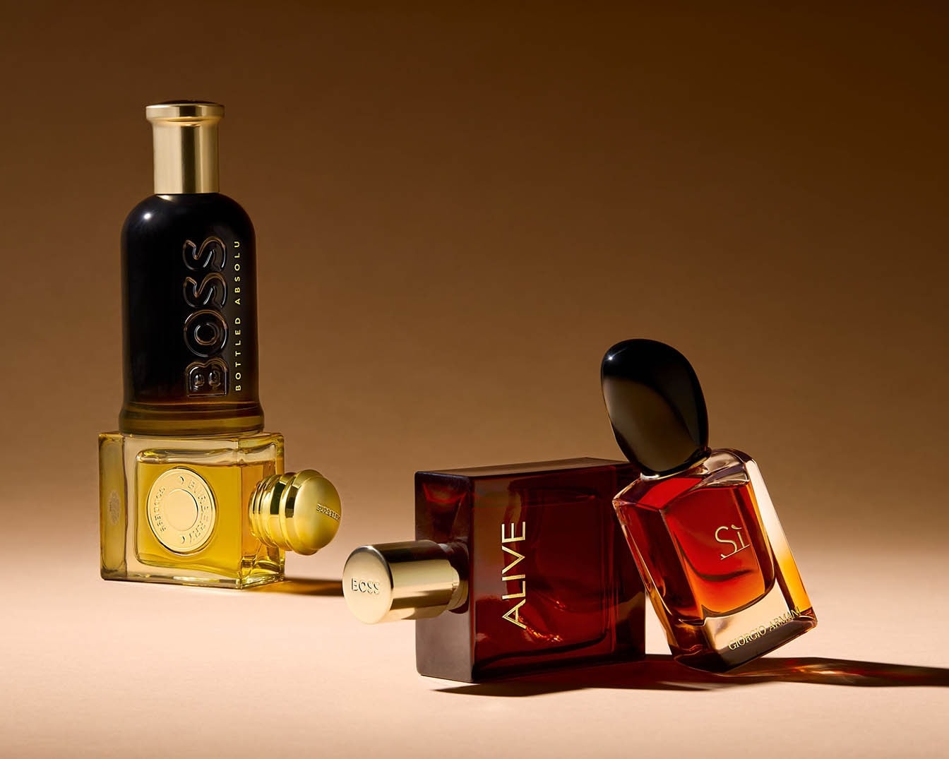 Drie parfumflessen, waaronder BOSS Bottled Absolu met een zwart-gouden flacon, ALIVE in een bruine flacon en Si Giorgio Armani met een rode flacon, elk met een unieke geur.