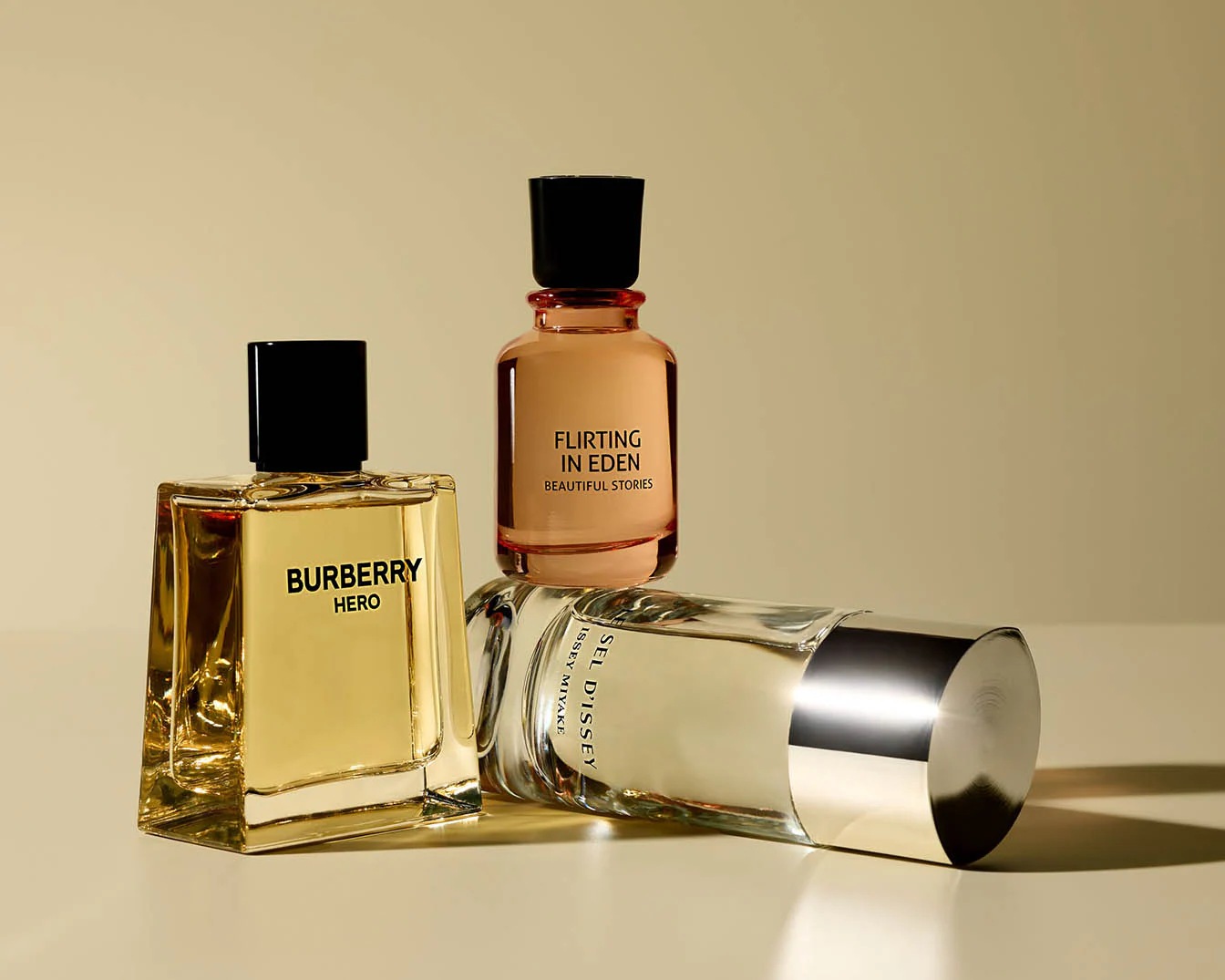 Drie parfumflessen staan op een rij: Burberry Hero met gouden vloeistof, Flirting in Eden met een perzikkleurige tint en Issey Miyake, liggend met een zilveren dop.