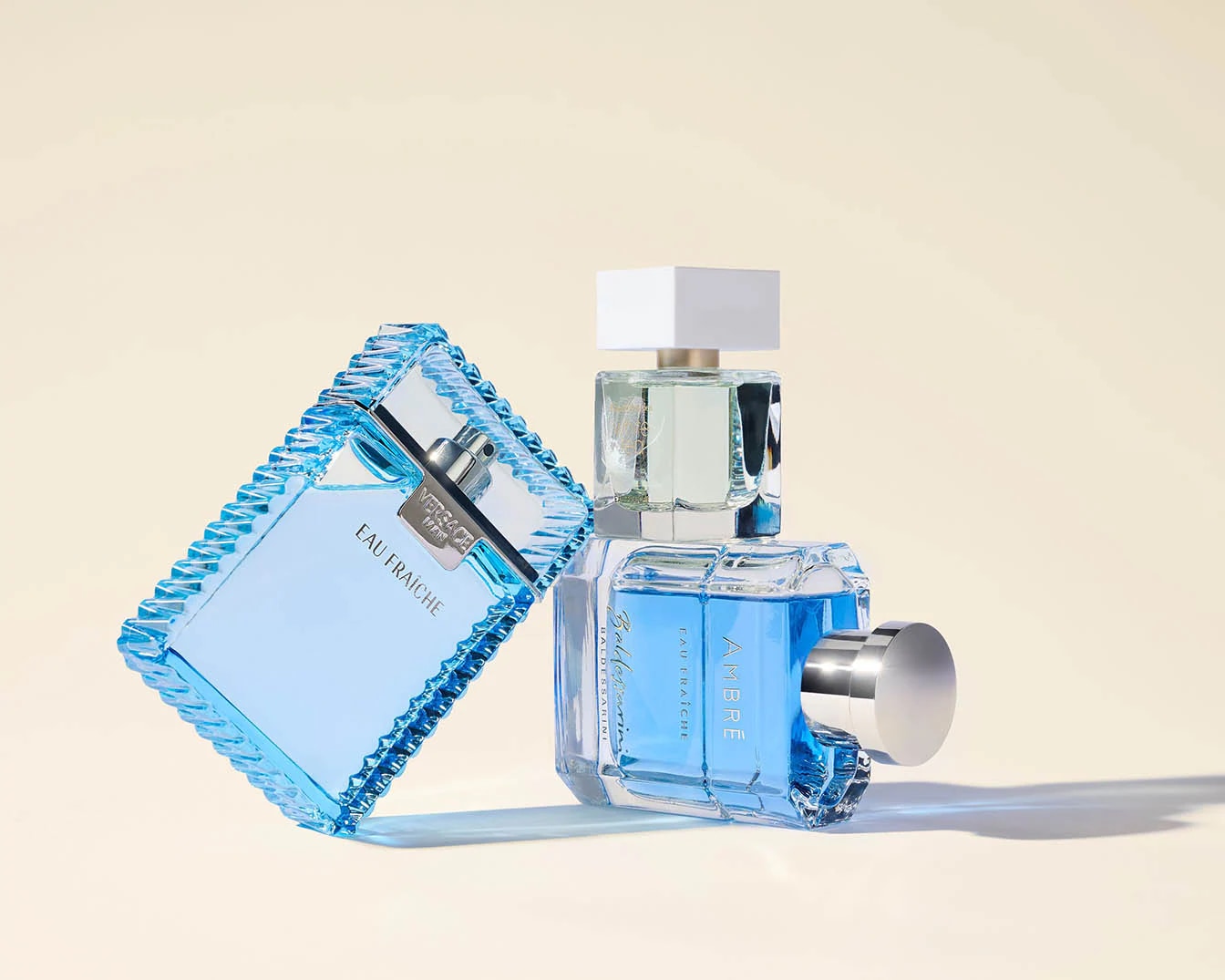 Twee blauwe parfumflessen, Versace Man Eau Fraiche met een helderblauwe vloeistof en Baldesarini Ambre Eau Fraiche met een diepblauwe vloeistof, staan op een zachte, crèmekleurige achtergrond.