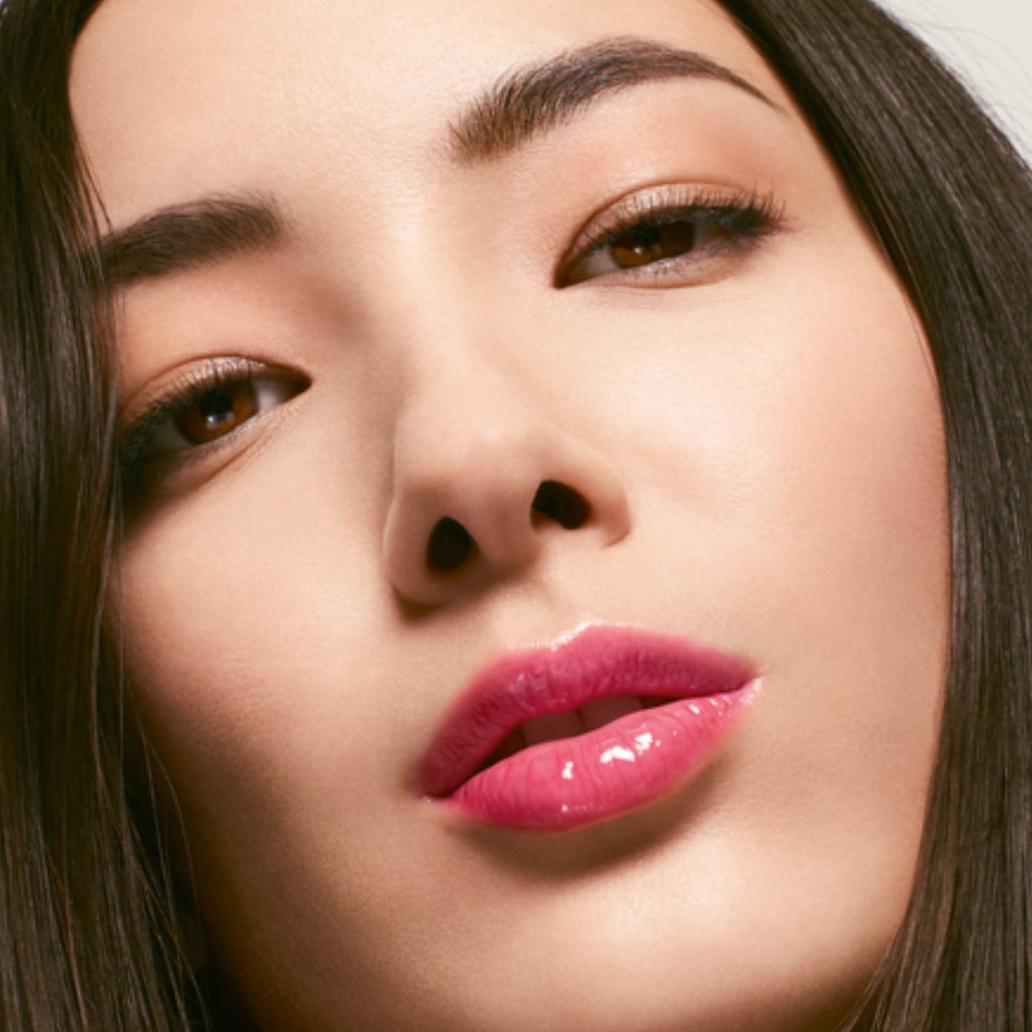 Gros plan sur un visage féminin avec des lèvres brillantes rose vif, des yeux marron doux et des sourcils bien définis, le tout encadré par des cheveux bruns lisses.