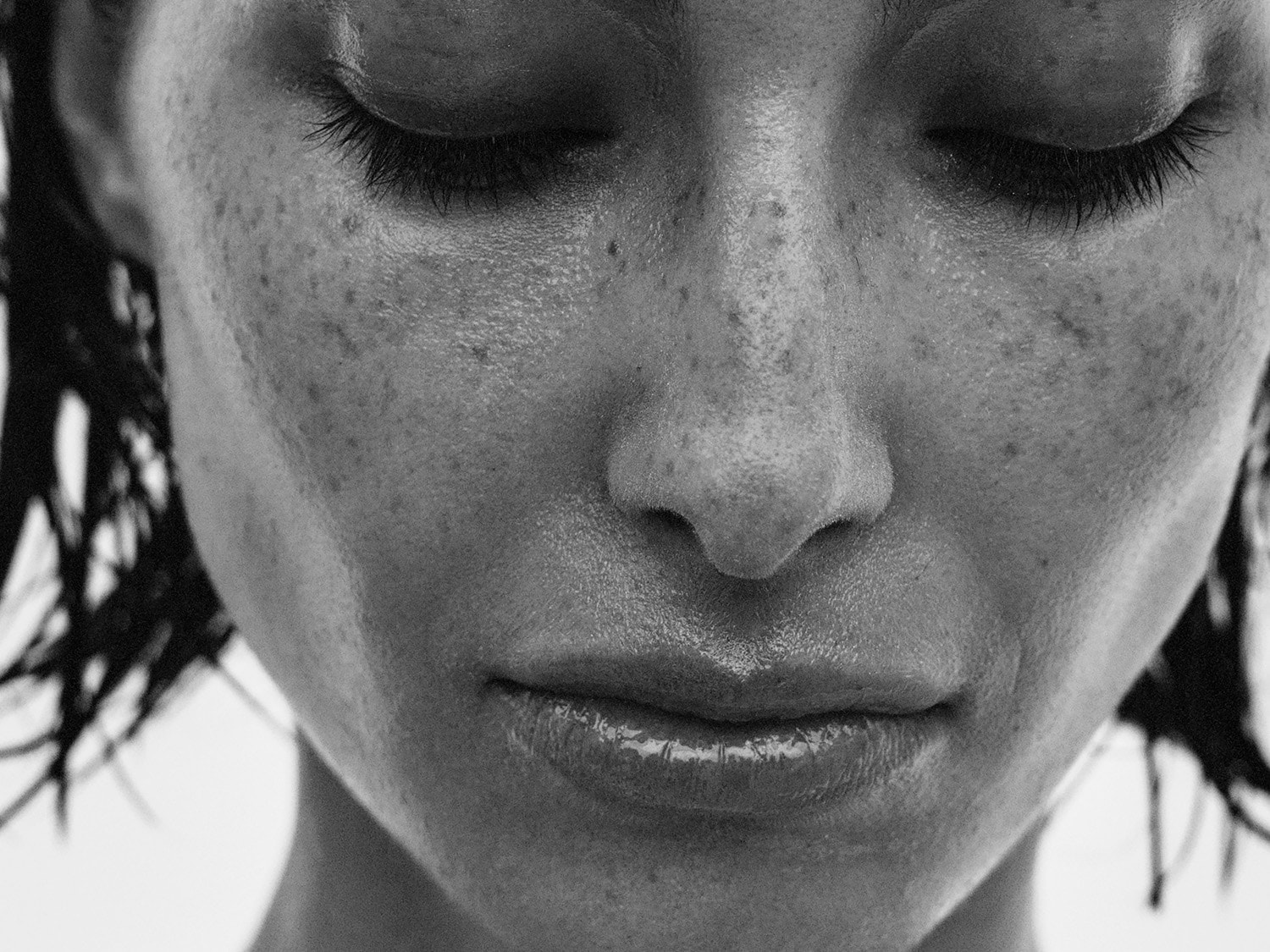 Juodai balta nuotrauka, kurioje matomas moters veidas su užmerktomis akimis, matomos strazdanos ant skruostų ir blizgančios lūpos.