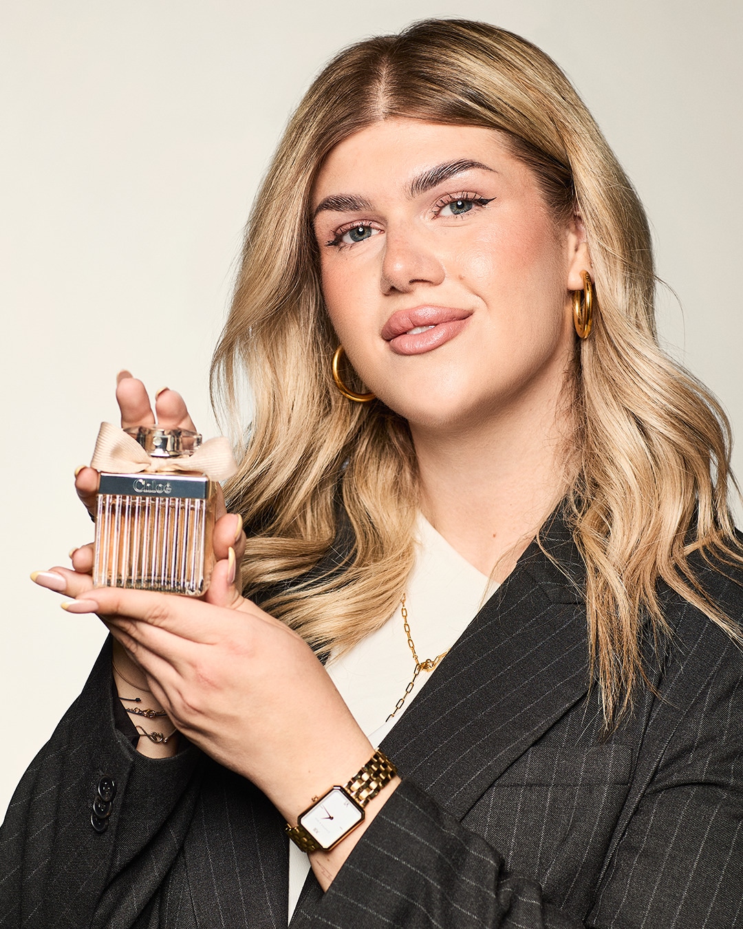 Eine blonde Frau hält ein Chloé-Parfum mit einer Schleife in den Händen und trägt einen Blazer, goldene Ohrringe und eine goldene Uhr.