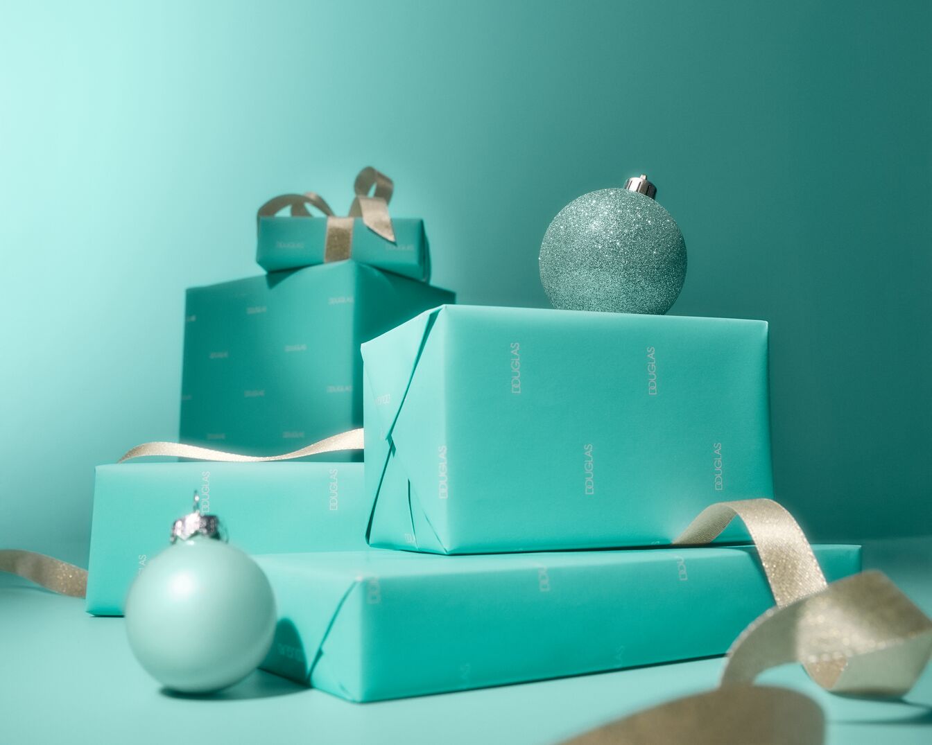 Na turkusowym tle piramida prezentów Douglas, przewiązanych złotymi wstążkami i ozdobionych błyszczącymi bombkami, tworzy świąteczny nastrój.