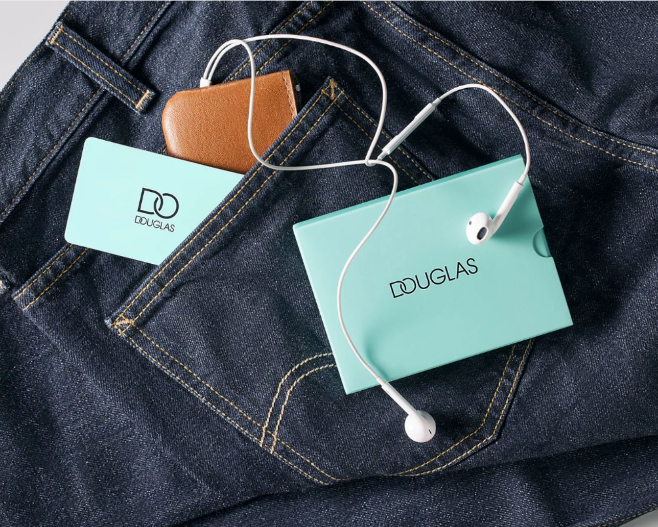 Na dżinsowym tle leżą słuchawki Douglas w turkusowym pudełku, brązowe etui i karta podarunkowa Douglas w kieszonce.