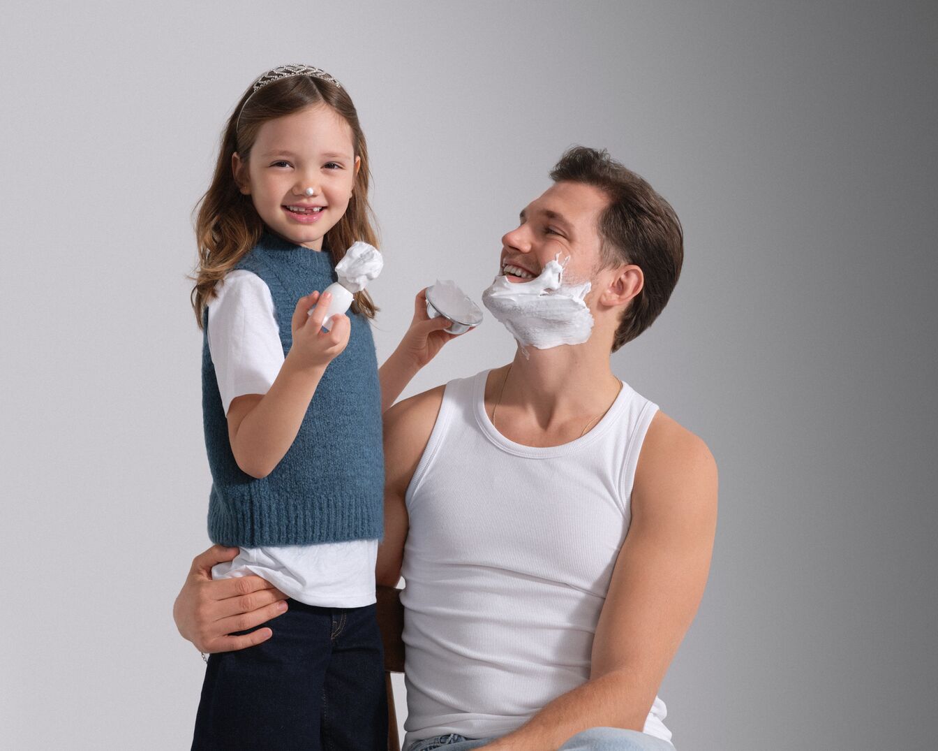 Uśmiechnięta dziewczynka w tiarze nakłada pędzlem piankę do golenia na twarz taty, który siedzi bez koszuli i śmieje się.
