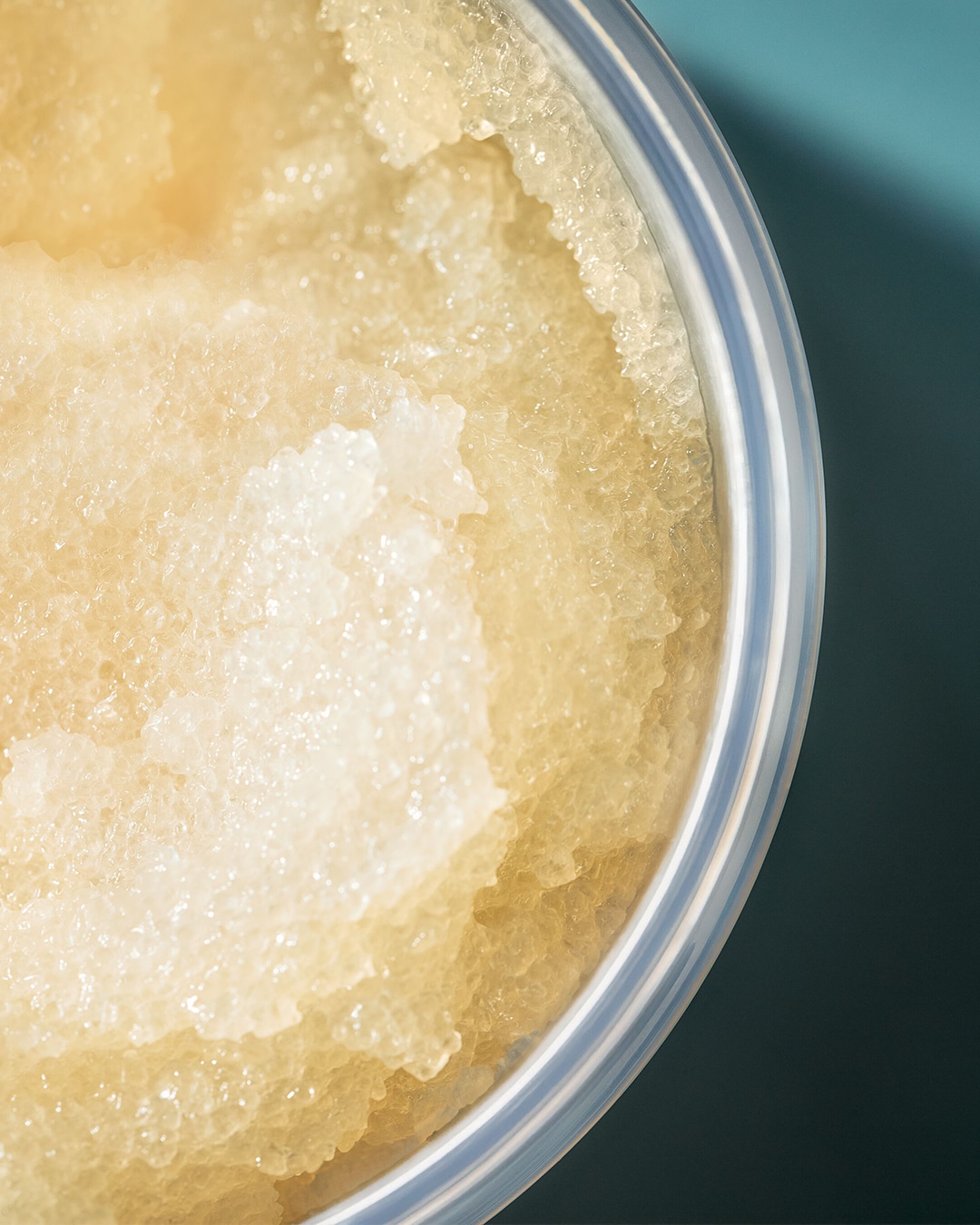 Nahaufnahme eines durchsichtigen Tiegels, gefüllt mit einem gelblichen Zuckerpeeling, das eine körnige Textur erkennen lässt.