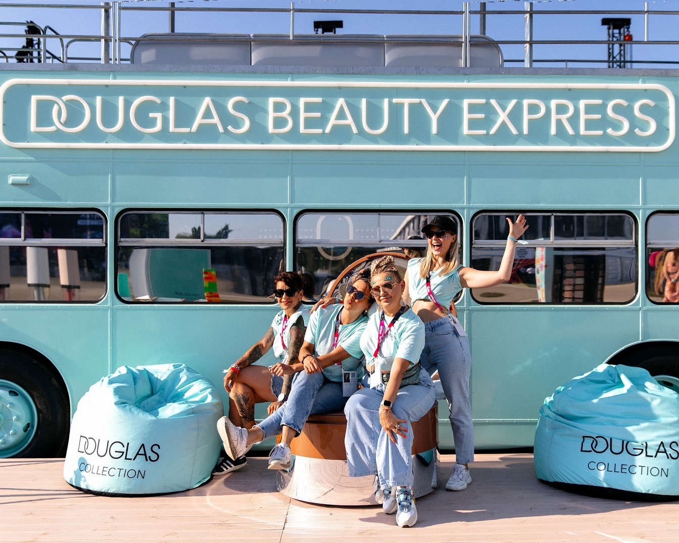 Cztery kobiety pozują na tle turkusowego autobusu Douglas Beauty Express, siedząc na krześle i pufach Douglas Collection.
