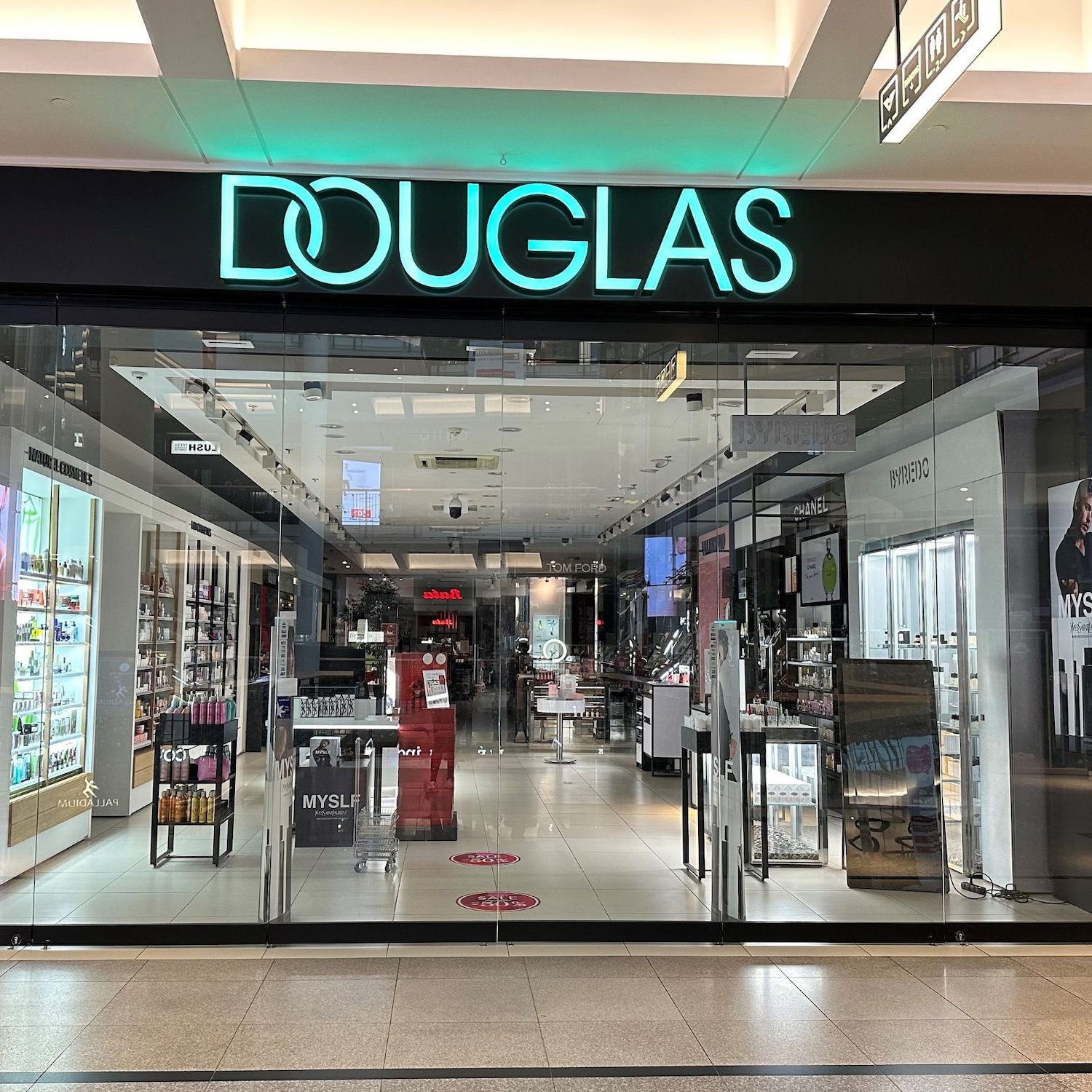 Pohled na vchod do prodejny Douglas s neonovým nápisem DOUGLAS nad skleněnými dveřmi, které odrážejí interiér plný kosmetiky a parfémů.
