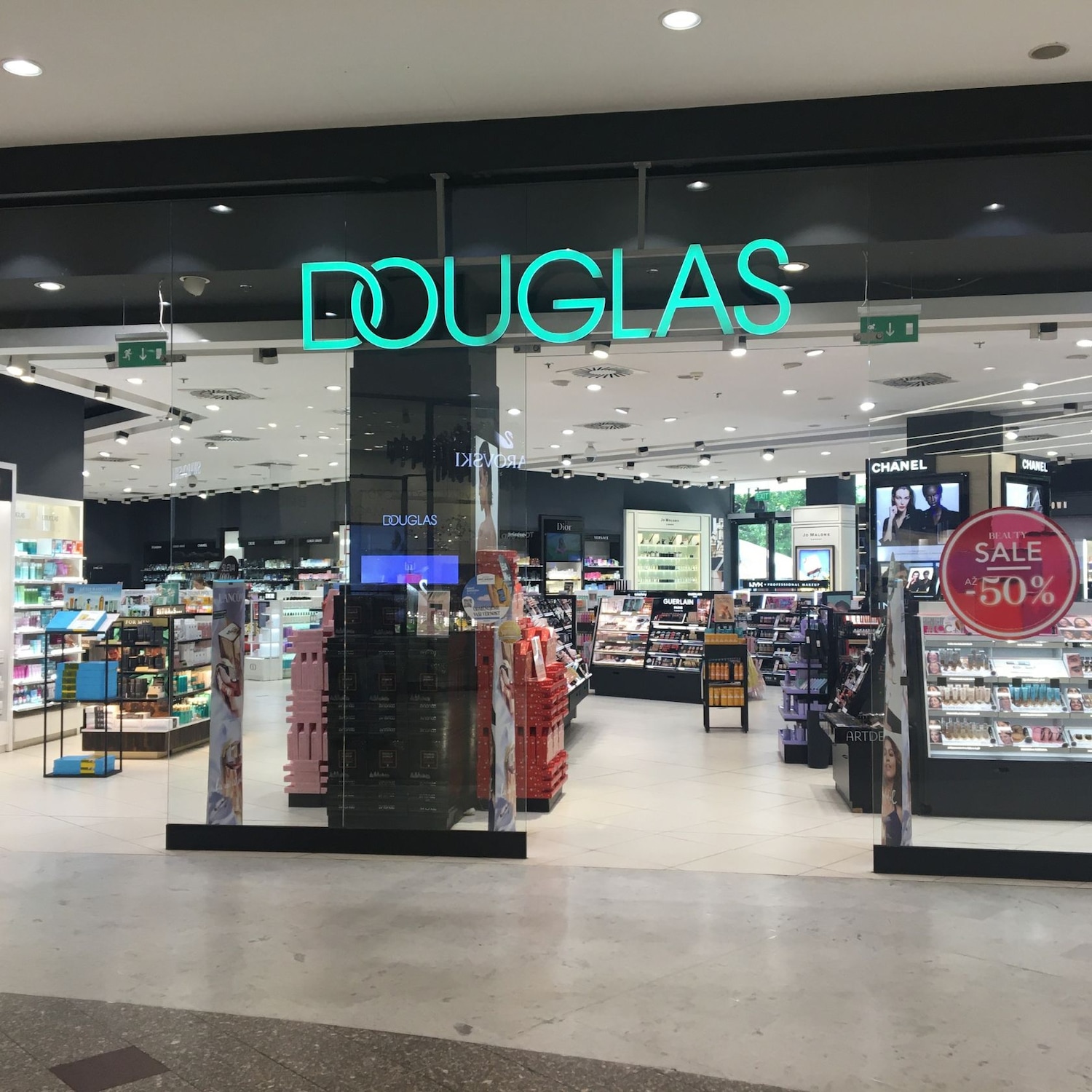 Pohled na prodejnu Douglas s nápisem Douglas v tyrkysové barvě nad vchodem, uvnitř viditelné regály s kosmetikou a akční nabídka Beauty Sale -50%.