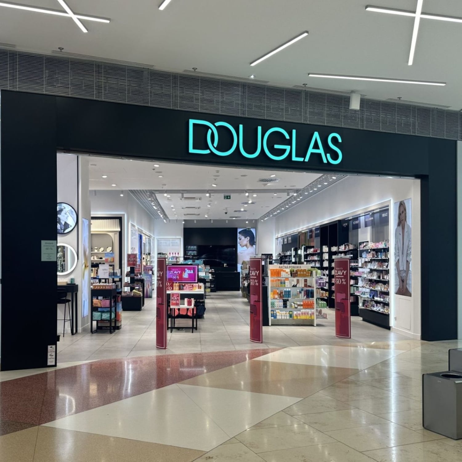 Vstup do predajne Douglas s modrým logom nad vchodom, ktorý vedie do dobre osvetleného interiéru s kozmetikou a parfumami.