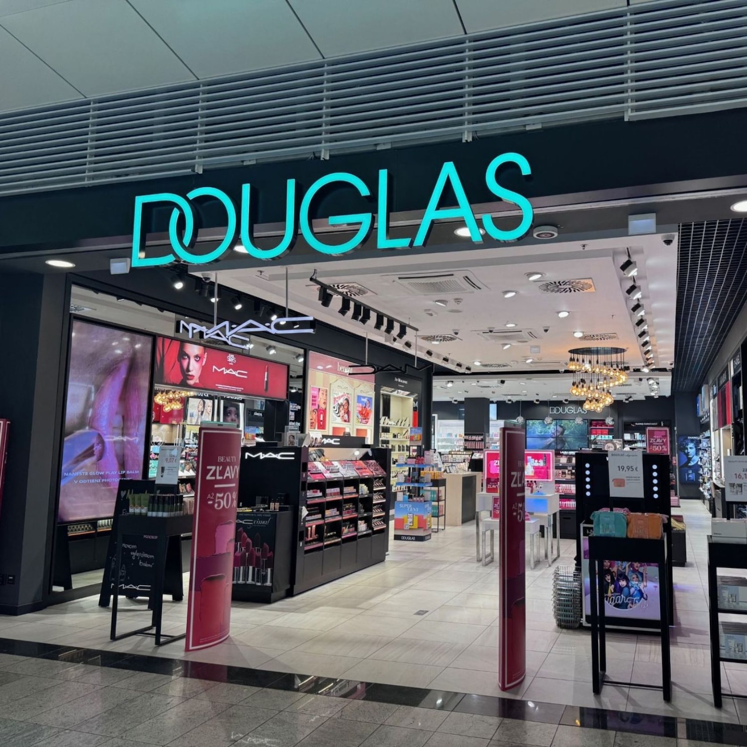 Pohľad na predajňu Douglas s výrazným neónovým logom a regálmi plnými kozmetiky značky MAC, ktoré lákajú na zľavy až do 50 %.