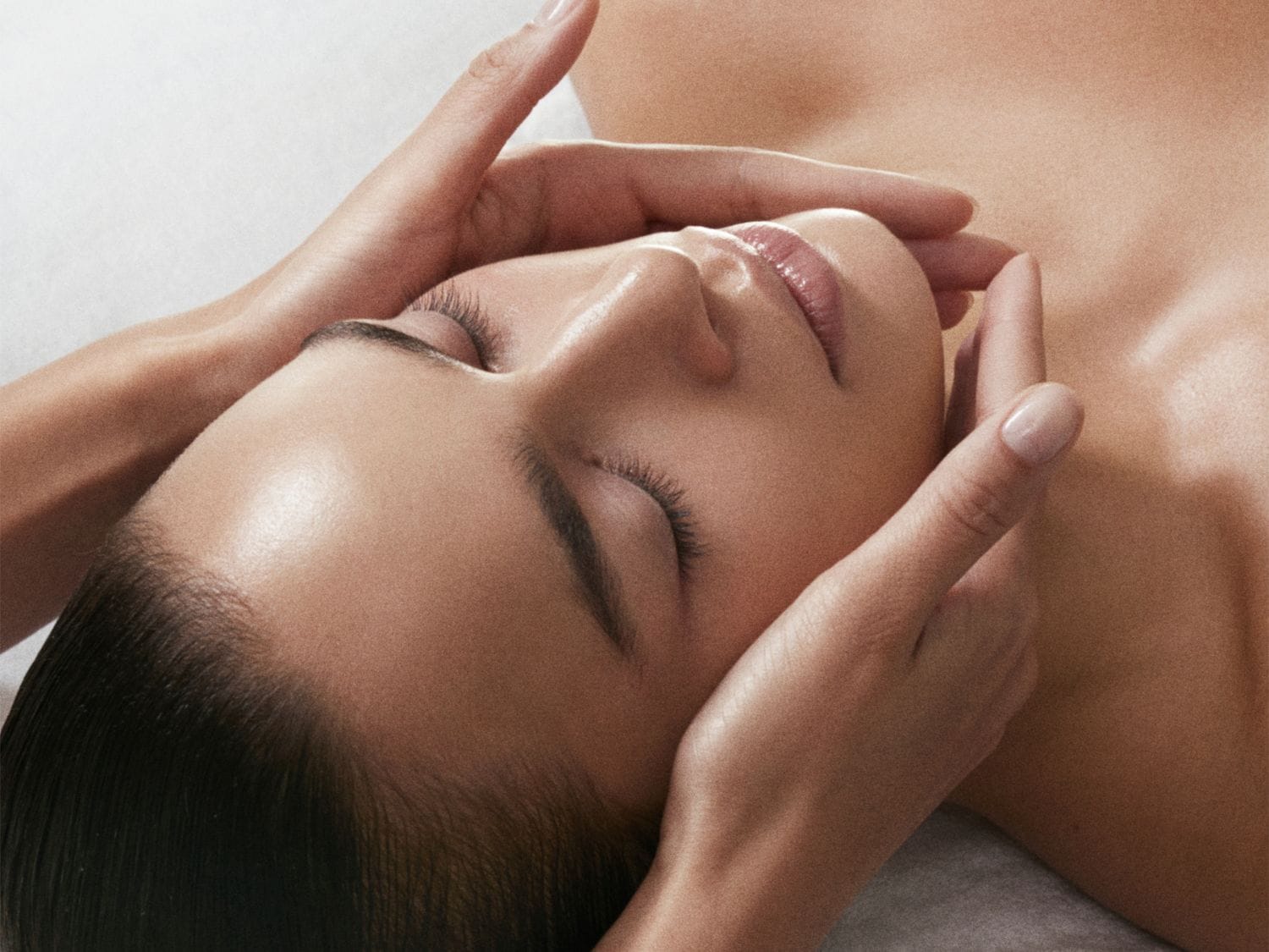 Egy nő arcmasszázst kap, a kezek finoman érintik az arcát, miközben ő csukott szemmel pihen.