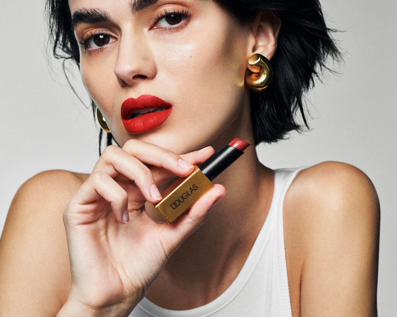 A modell élénkpiros rúzzsal, arany fülbevalóval és egy Douglas márkájú, arany színű rúzsos tubussal pózol.