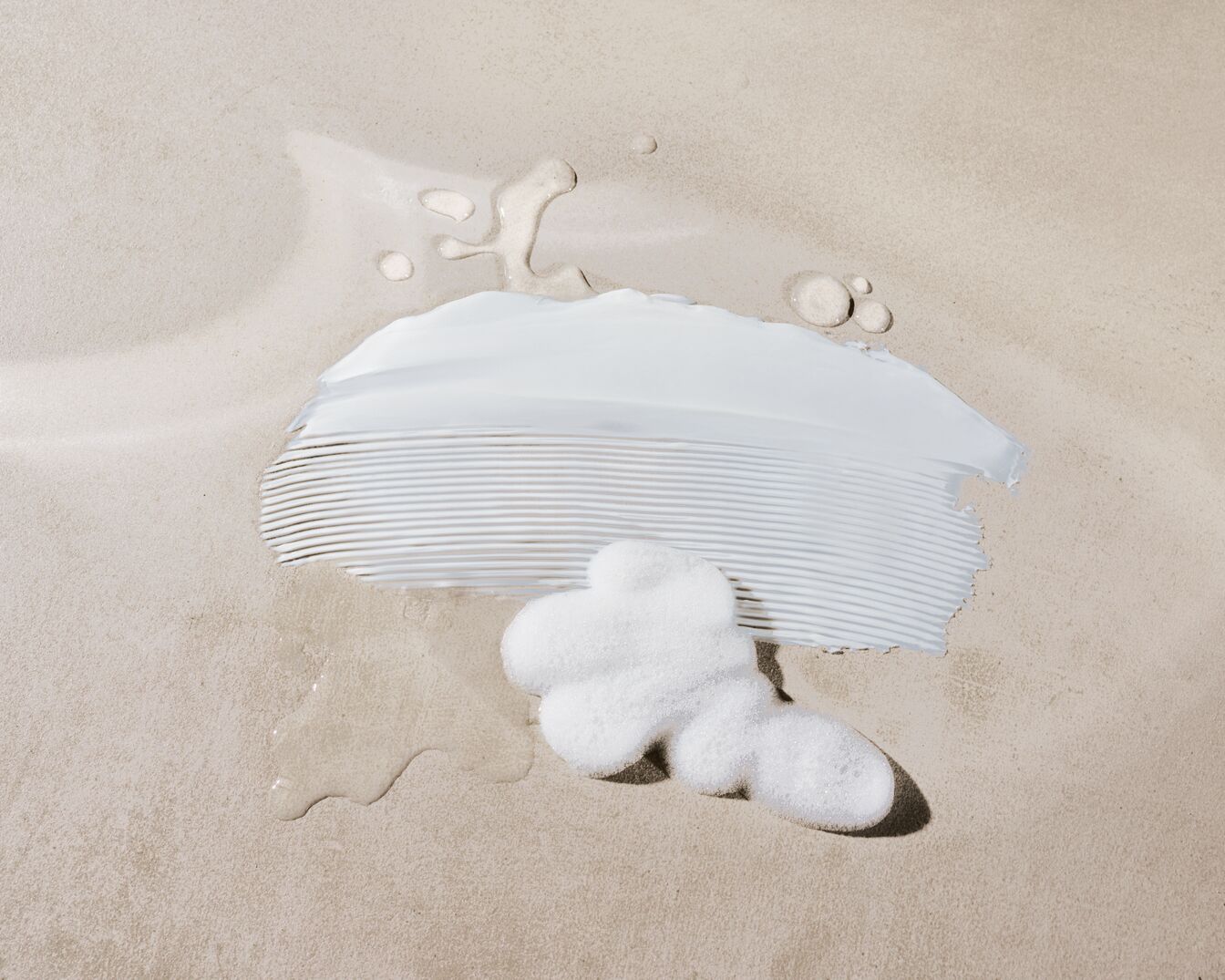 Nahaufnahme von weißer Creme, schaumigem Badesalz und Wassertropfen auf beigem Untergrund, die die Textur und Feuchtigkeit der Hautpflegeprodukte hervorheben.