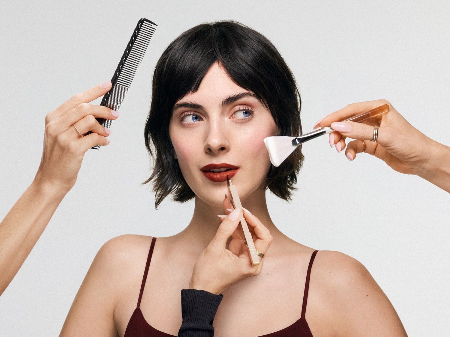 Na obrázku je žena s krátkymi tmavými vlasmi, na ktorej sa vykonáva make-up pomocou štetcov, ceruzky na pery a hrebeňa.