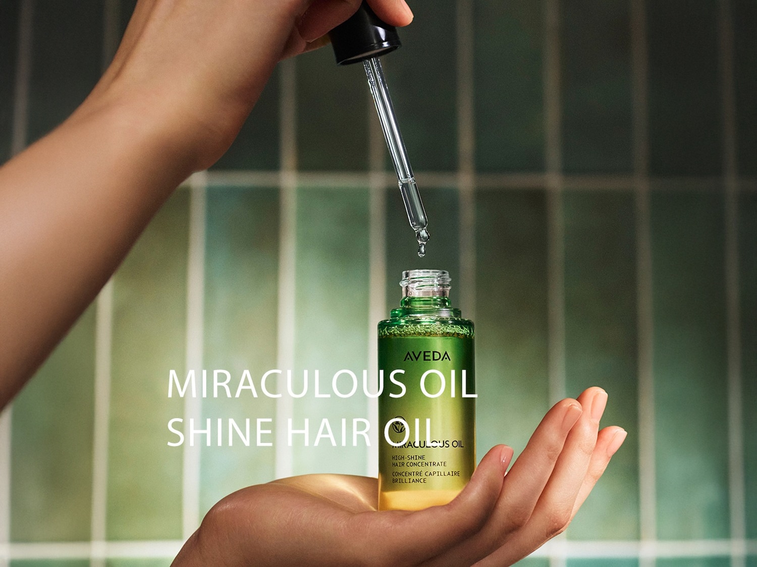 Een hand houdt een flesje Aveda Miraculous Oil Shine Hair Oil vast, terwijl een pipet een druppel olie laat vallen, wat de hoogglans haarconcentraat benadrukt.