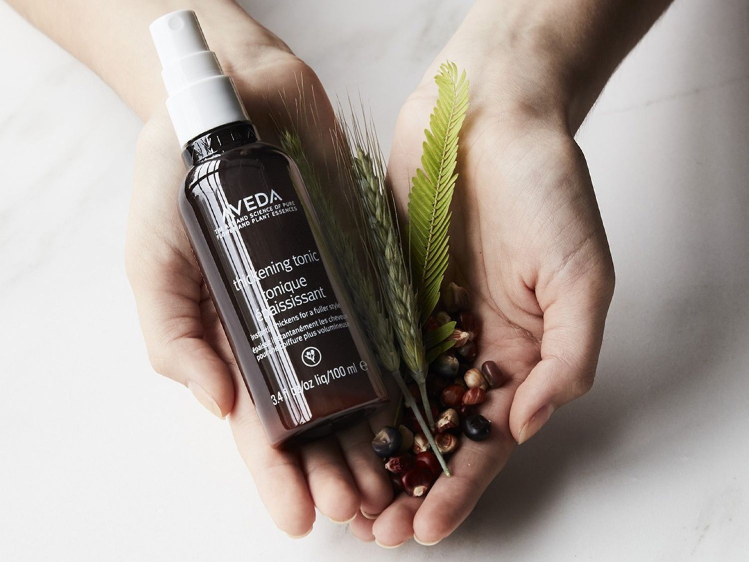 Twee handen houden een bruine fles Aveda Thickening Tonic vast, samen met tarwe en zaden, voor een vollere haarstijl.