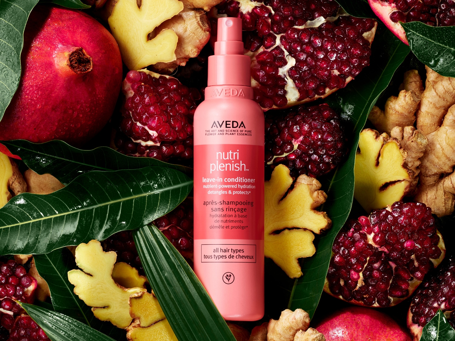 Een roze fles Aveda Nutriplenish leave-in conditioner, omringd door granaatappels, gember en groene bladeren, voor hydratatie en bescherming van alle haartypes.