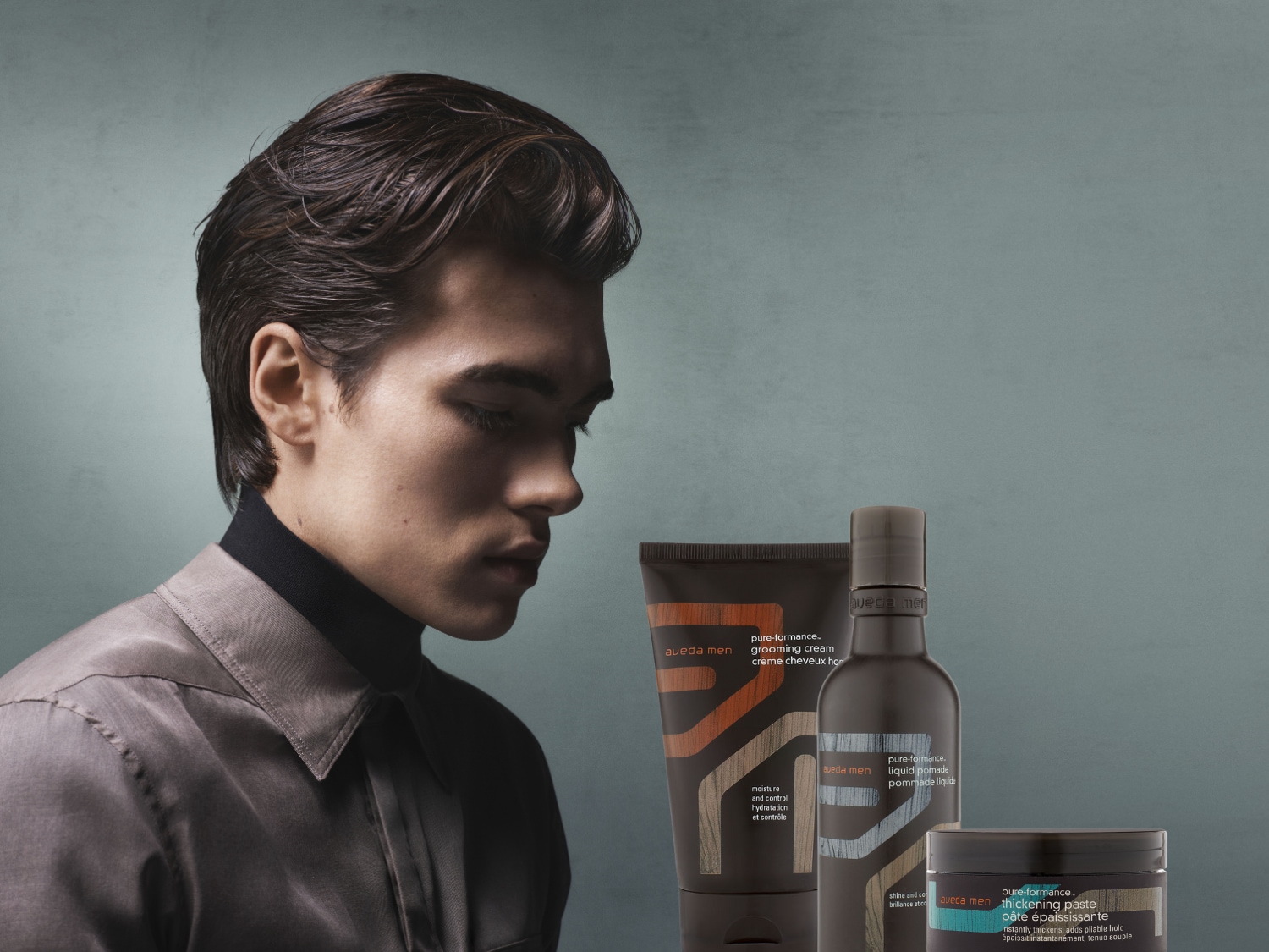 Een man met gestyled haar staat naast Aveda Men haarproducten, waaronder grooming cream, liquid pomade en thickening paste, voor hydratatie, controle en glans.