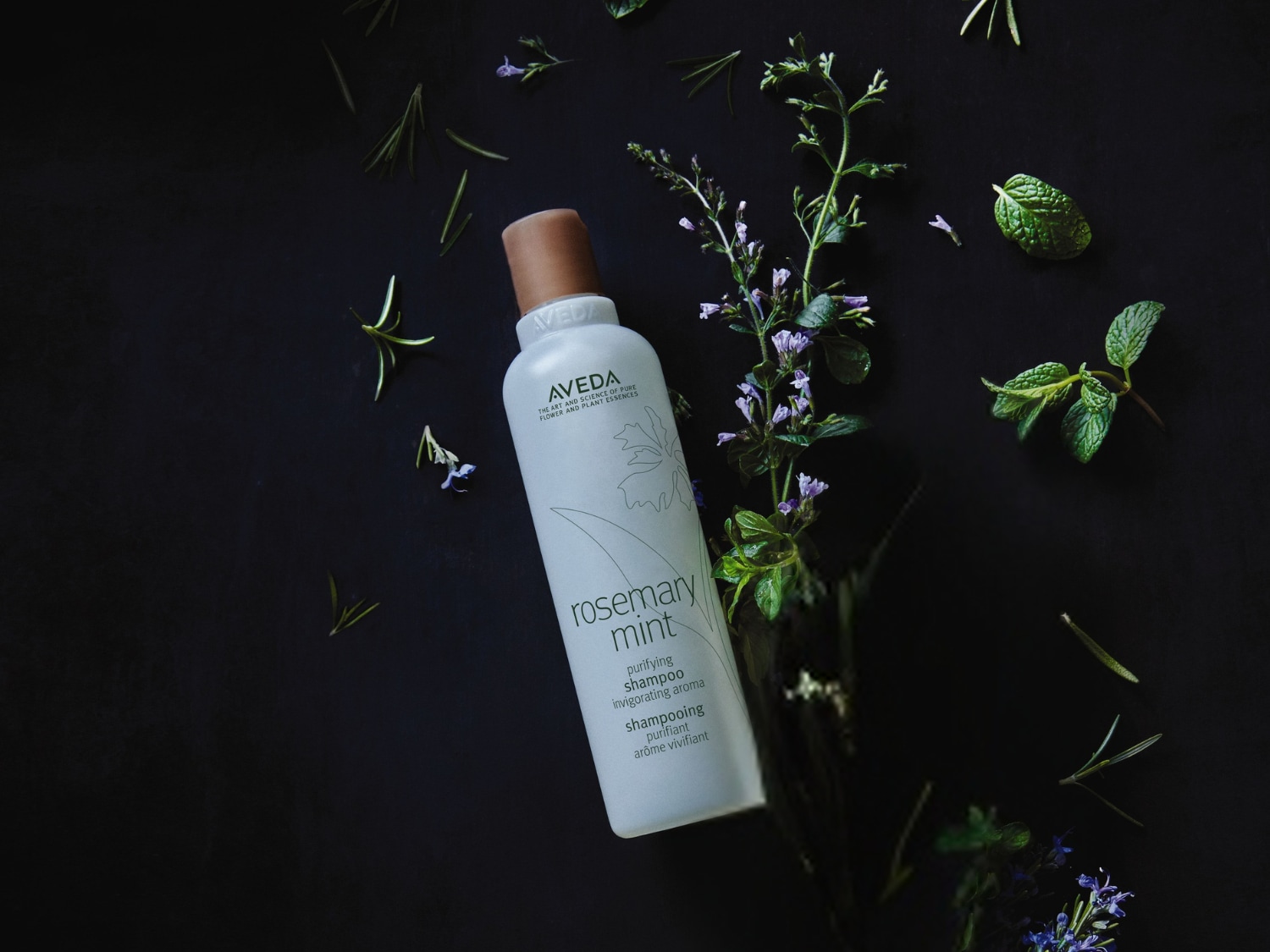 De Aveda Rosemary Mint zuiverende shampoo in een witte fles met bruine dop, omringd door rozemarijn en munt, voor een verkwikkende geur.