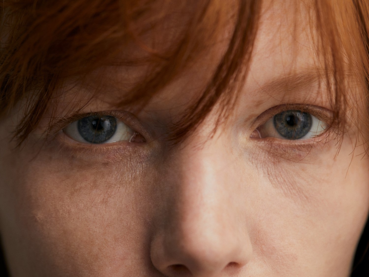 Nahaufnahme von einem Gesicht mit hellblauen Augen und roten Haaren, die Stirn bedecken.