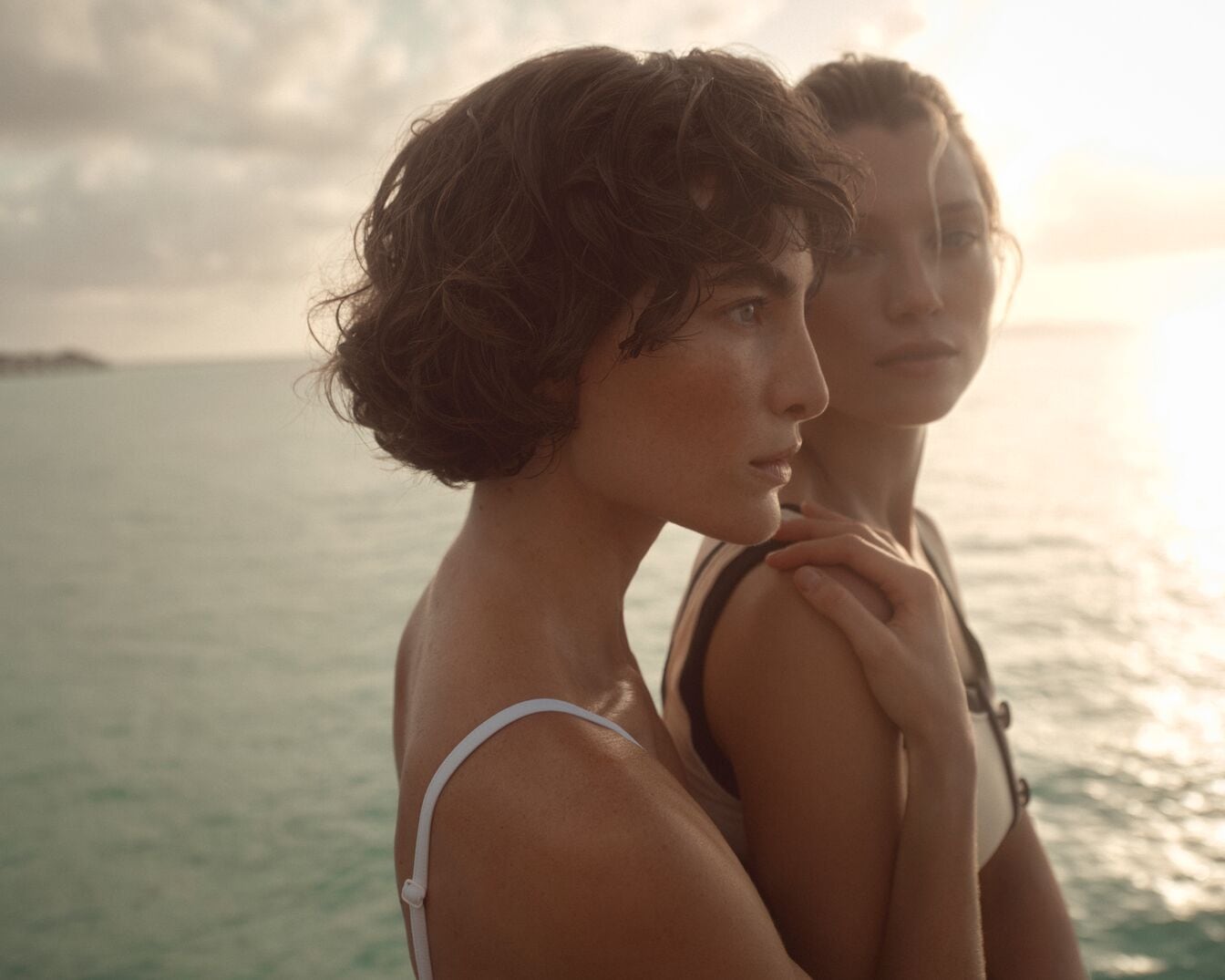 Zwei Frauen stehen vor dem Hintergrund des Meeres und des Himmels, wobei eine Frau die Schulter der anderen berührt, was eine Atmosphäre von Intimität und Verbundenheit erzeugt.