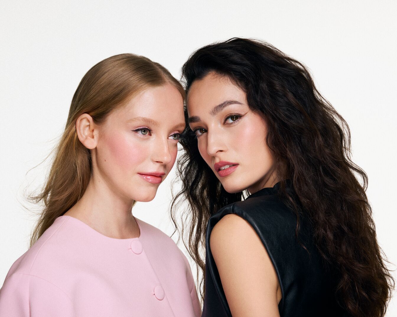 Zwei Frauen, eine mit hellbraunem, glattem Haar und rosa Blazer, die andere mit gewelltem, dunklem Haar und schwarzem Top, beide mit dezentem Make-up.