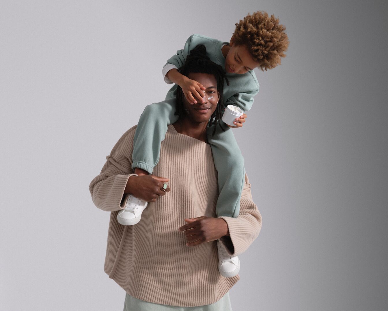 Un enfant assis sur les épaules d'un homme lui met de la crème sur le visage, tenant un pot blanc dans sa main.