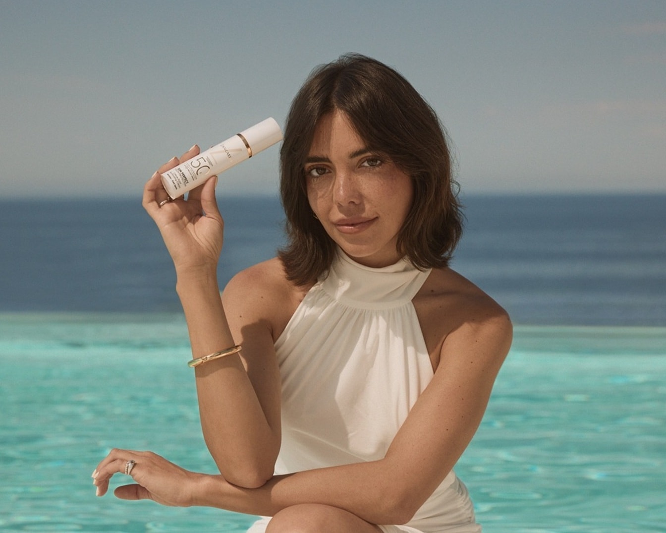 Una mujer sostiene un protector solar Lancome con SPF 50 en un envase blanco y dorado, posando con un vestido blanco y el mar azul de fondo.