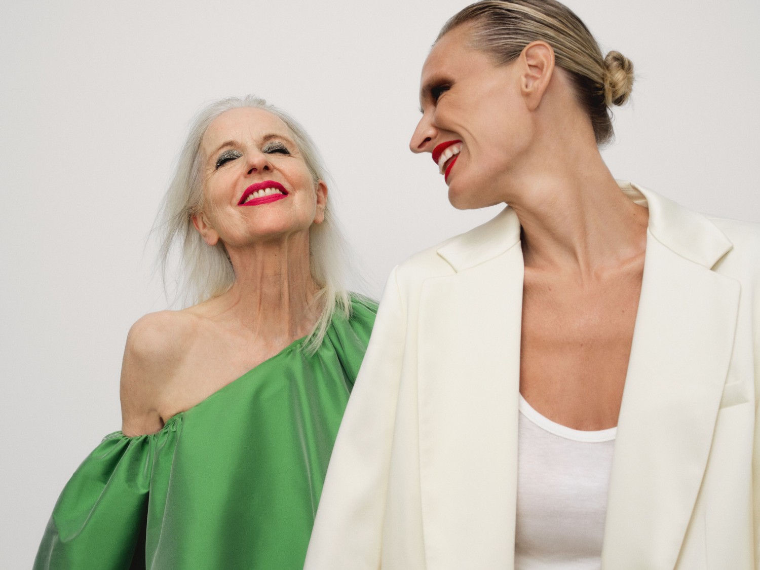 Švytinčios moterys su raudonais lūpų dažais pozuoja: viena su žalia suknele atidengtais pečiais, kita su kremine palaidine ir švarku.