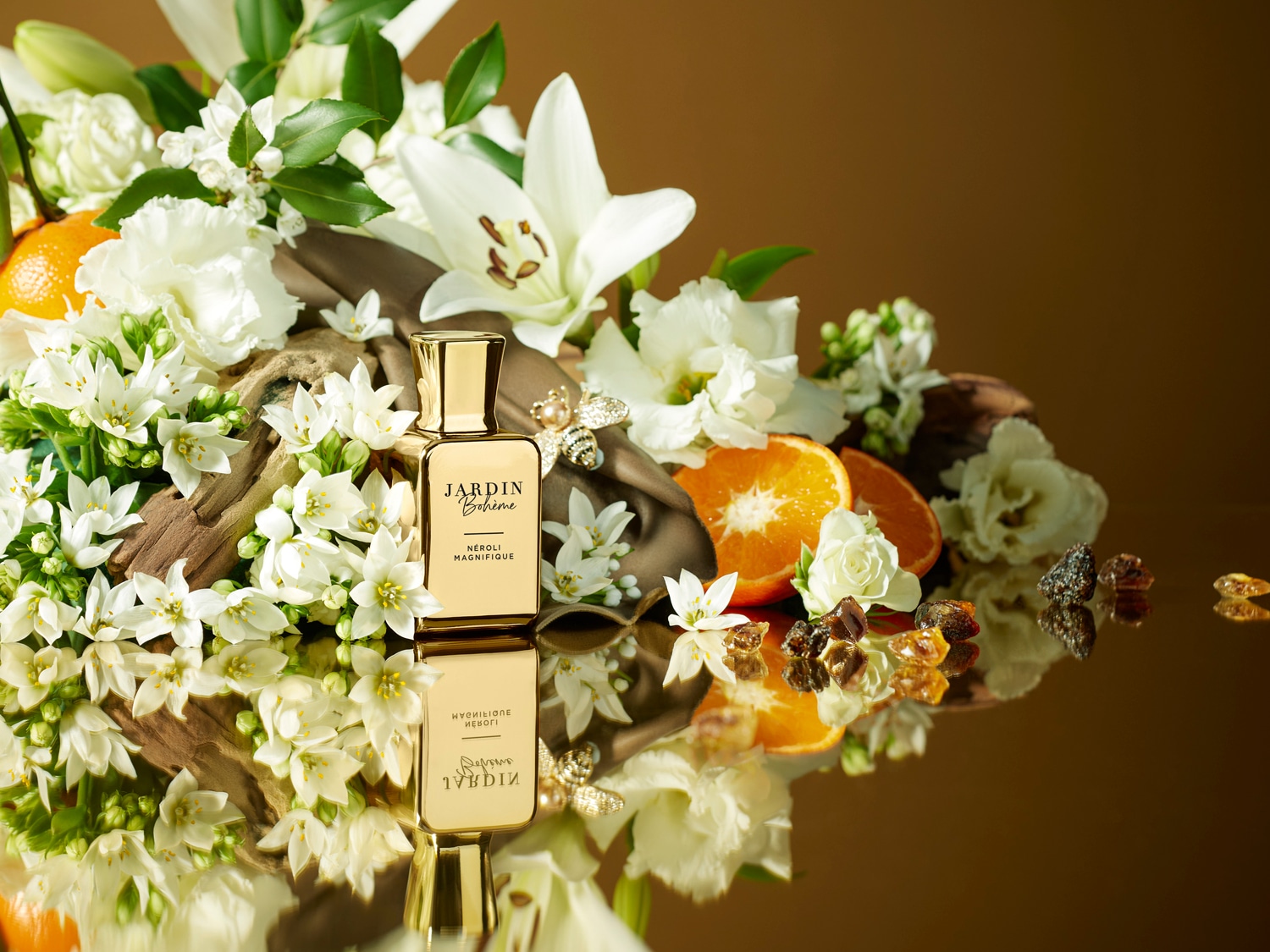 Auksinis „Jardin Bohème Néroli Magnifique“ kvepalų buteliukas, apsuptas baltų gėlių, apelsinų skiltelių ir gintaro spalvos akmenukų, atspindi prabangą ir gamtos grožį.