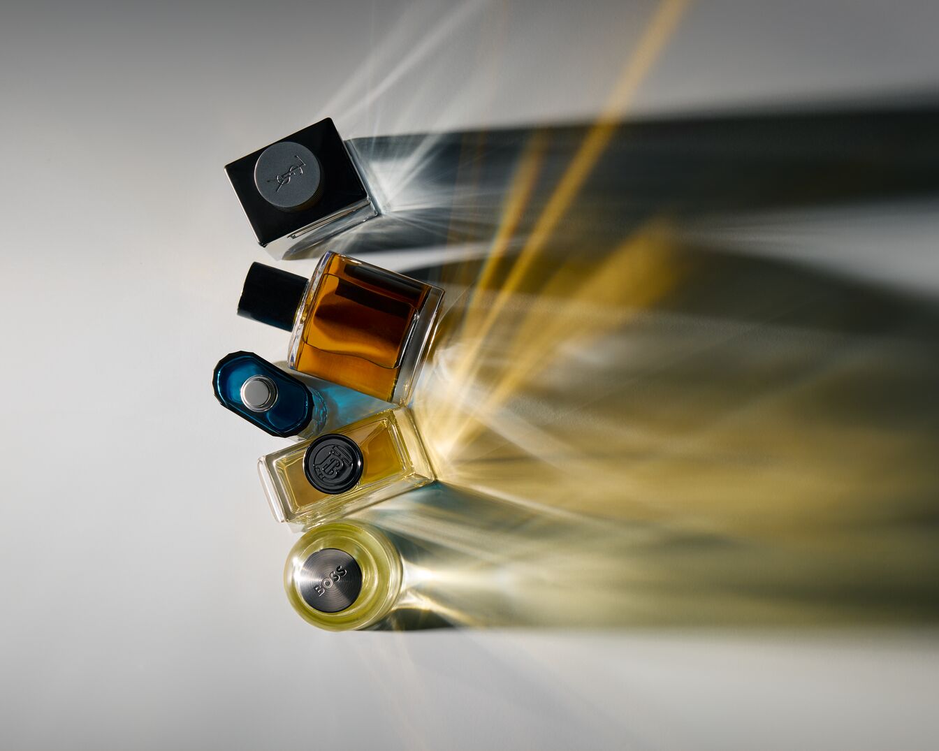 Įvairių formų ir spalvų kvepalų buteliukai išdėstyti ant balto paviršiaus, apšviesti ryškia šviesa, sukuriančia ilgesnius šešėlius.