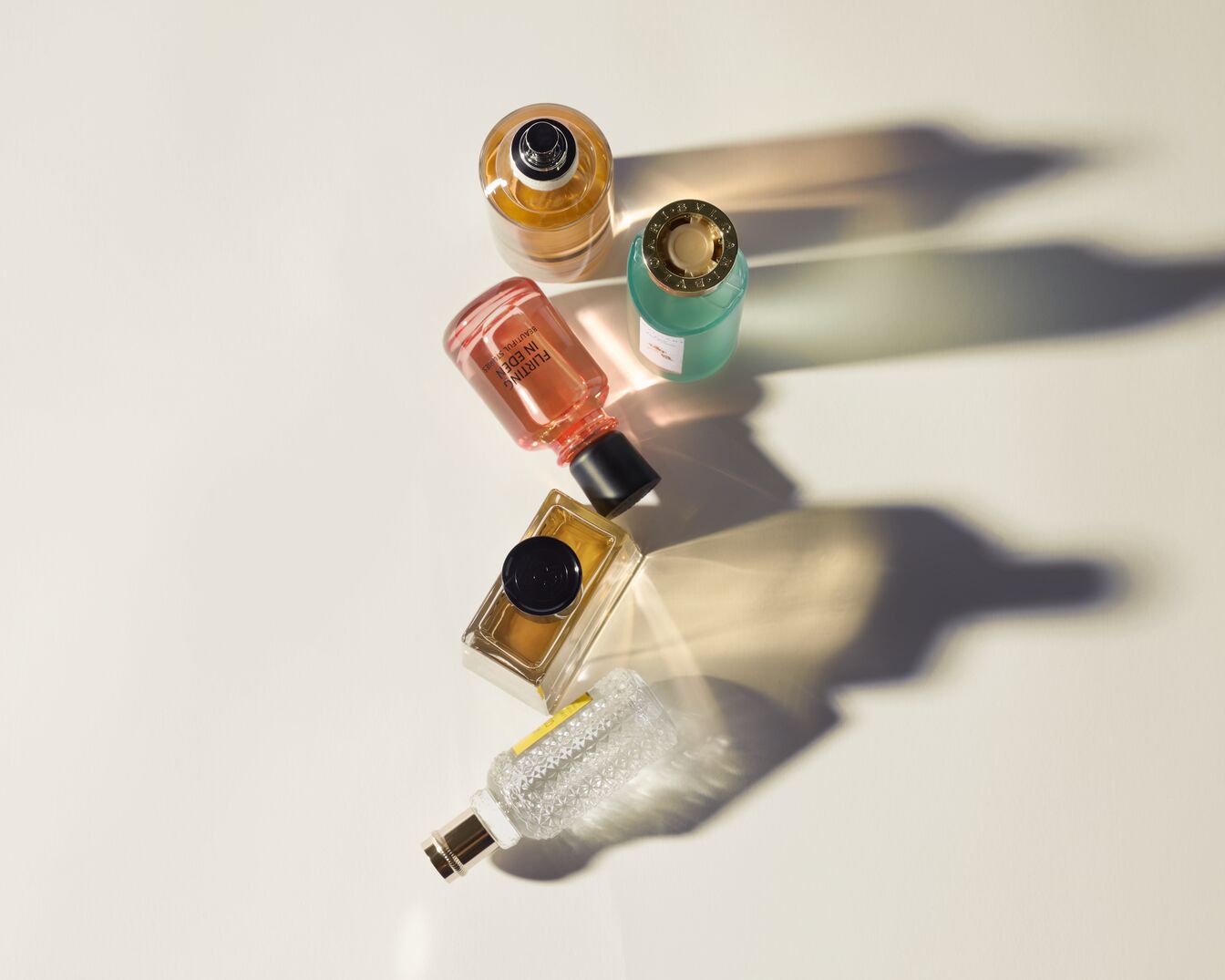 Įvairių formų ir spalvų kvepalų buteliukai išdėstyti ant balto paviršiaus, sukuria žaismingą kompoziciją.