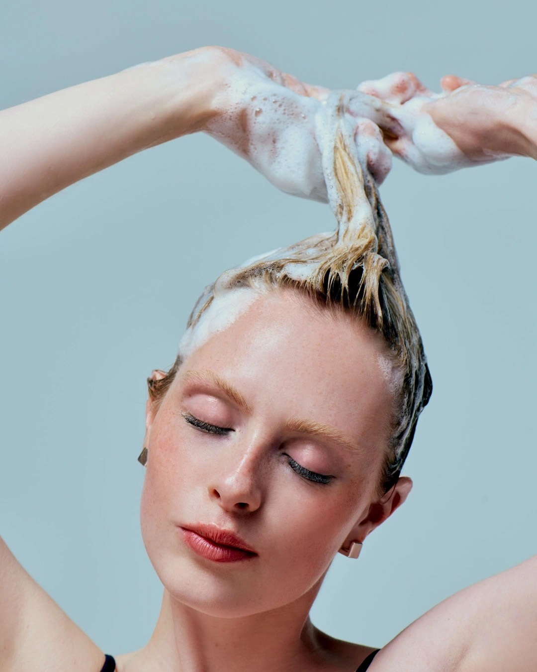 Een vrouw met gesloten ogen wast haar blonde haar met schuimende shampoo.