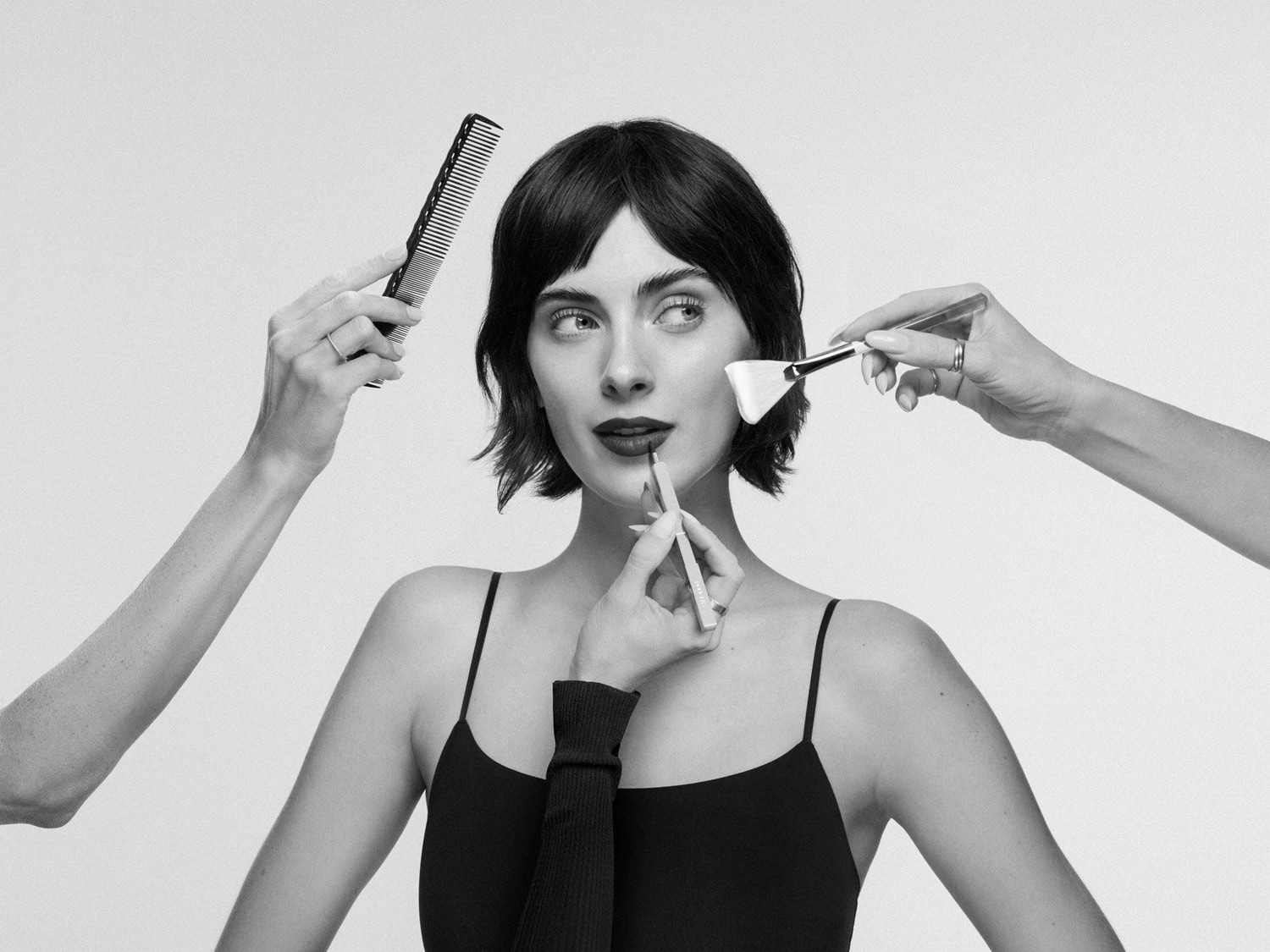 Eine Frau mit dunklen Haaren und dunklem Lippenstift wird von mehreren Händen mit einem Kamm, einem Lippenstift und einem Pinsel geschminkt.