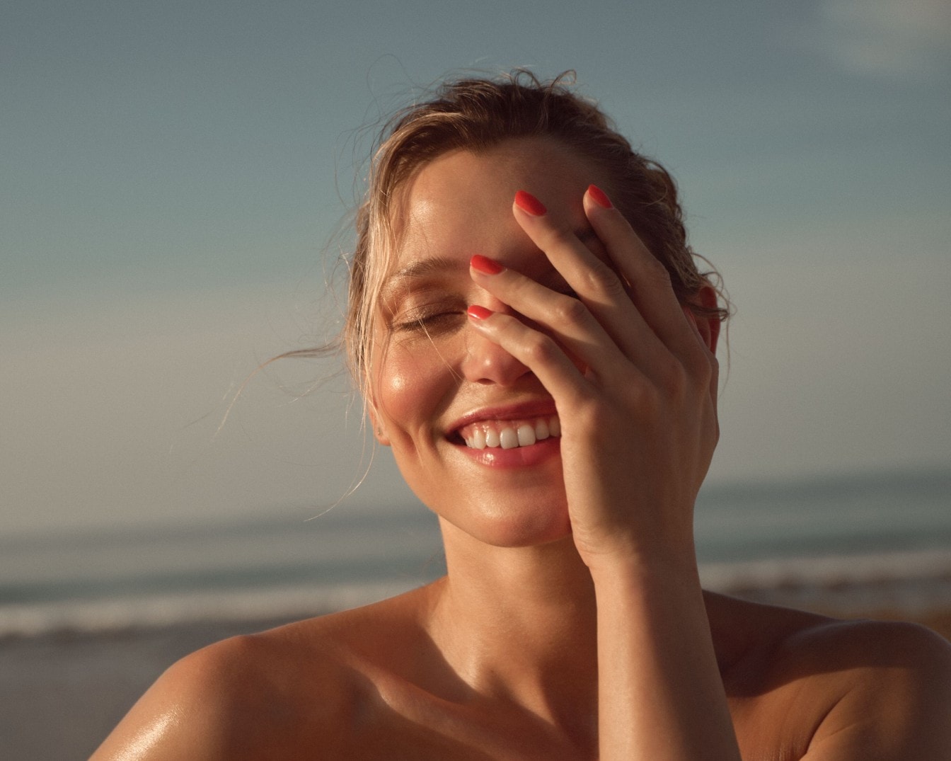 Eine strahlende Frau mit roten Nägeln lächelt, während sie ihre Augen mit der Hand vor der Sonne schützt, mit einem Hintergrund aus Meer und Himmel.