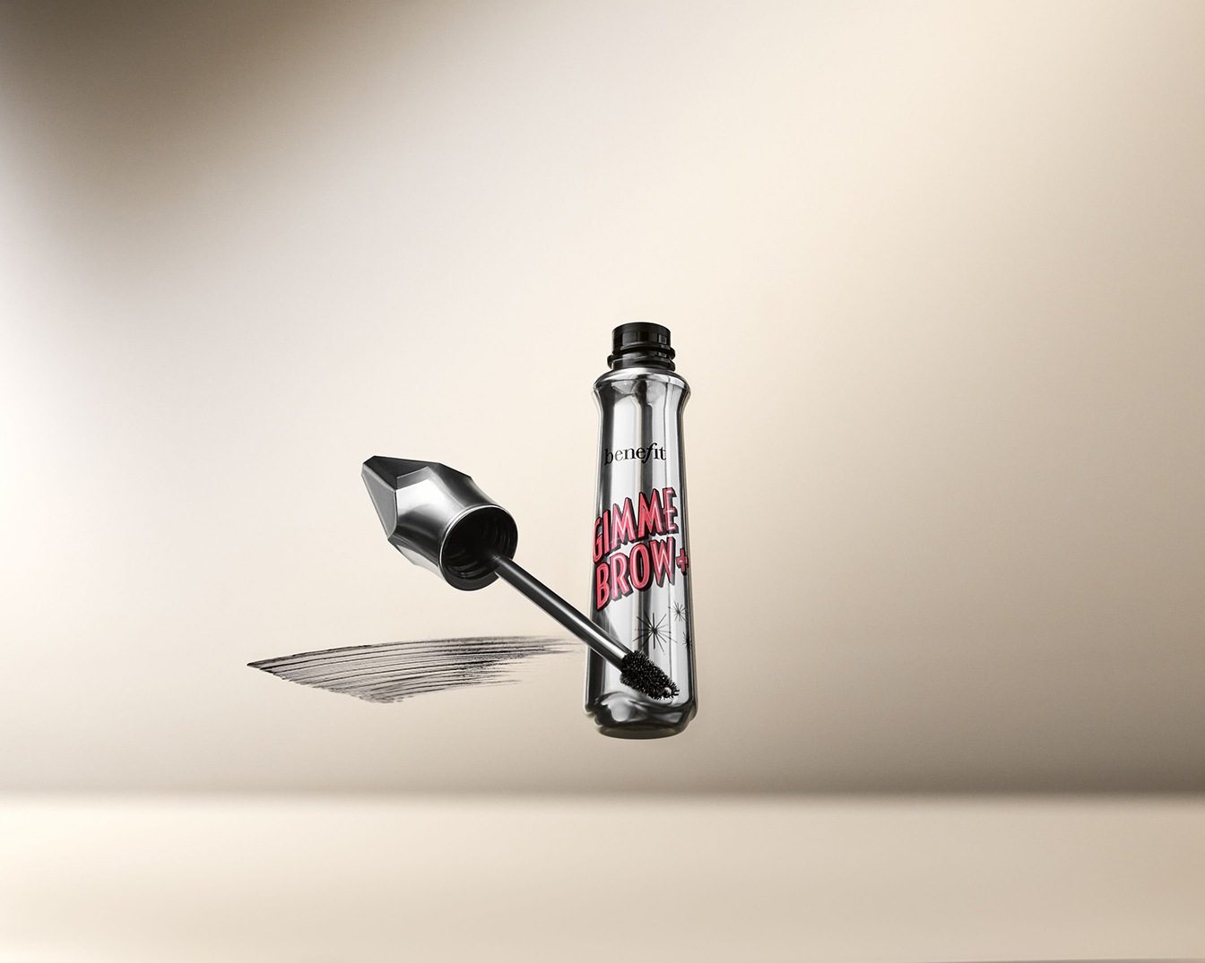 Zilveren Benefit Gimme Brow+ wenkbrauwgel met een zwarte dop en roze letters, de borstel ligt ernaast met een veeg van de gel.