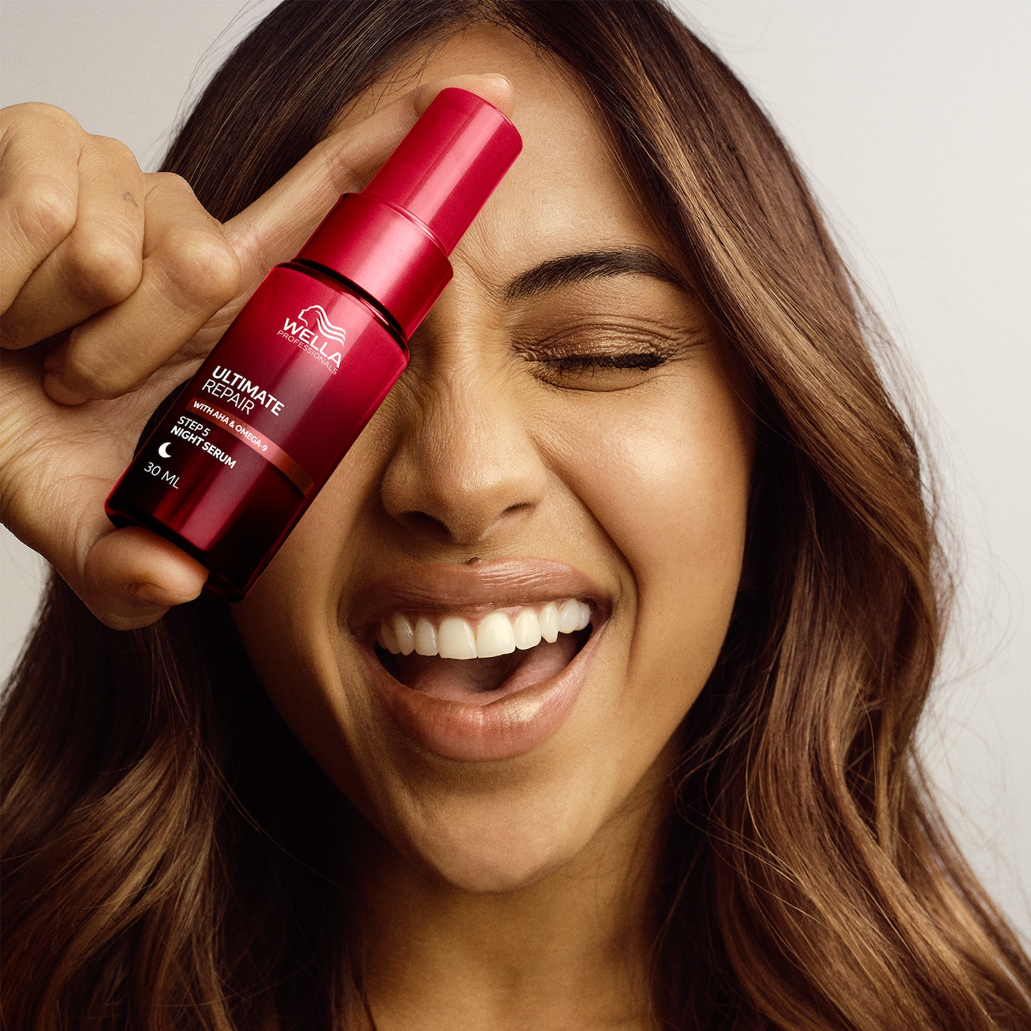 Una donna sorridente tiene in mano il siero notte Ultimate Repair di Wella Professionals, confezionato in un flacone rosso scuro da 30 ml.