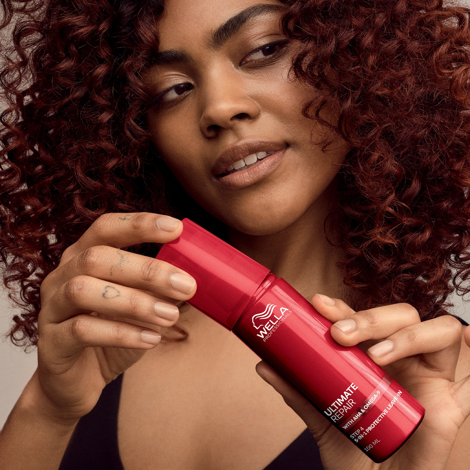 Una donna dai capelli ricci castano-rossicci tiene in mano un flacone rosso di Wella Professionals Ultimate Repair, un leave-in protettivo 5 in 1 con AHA e Omega-9.
