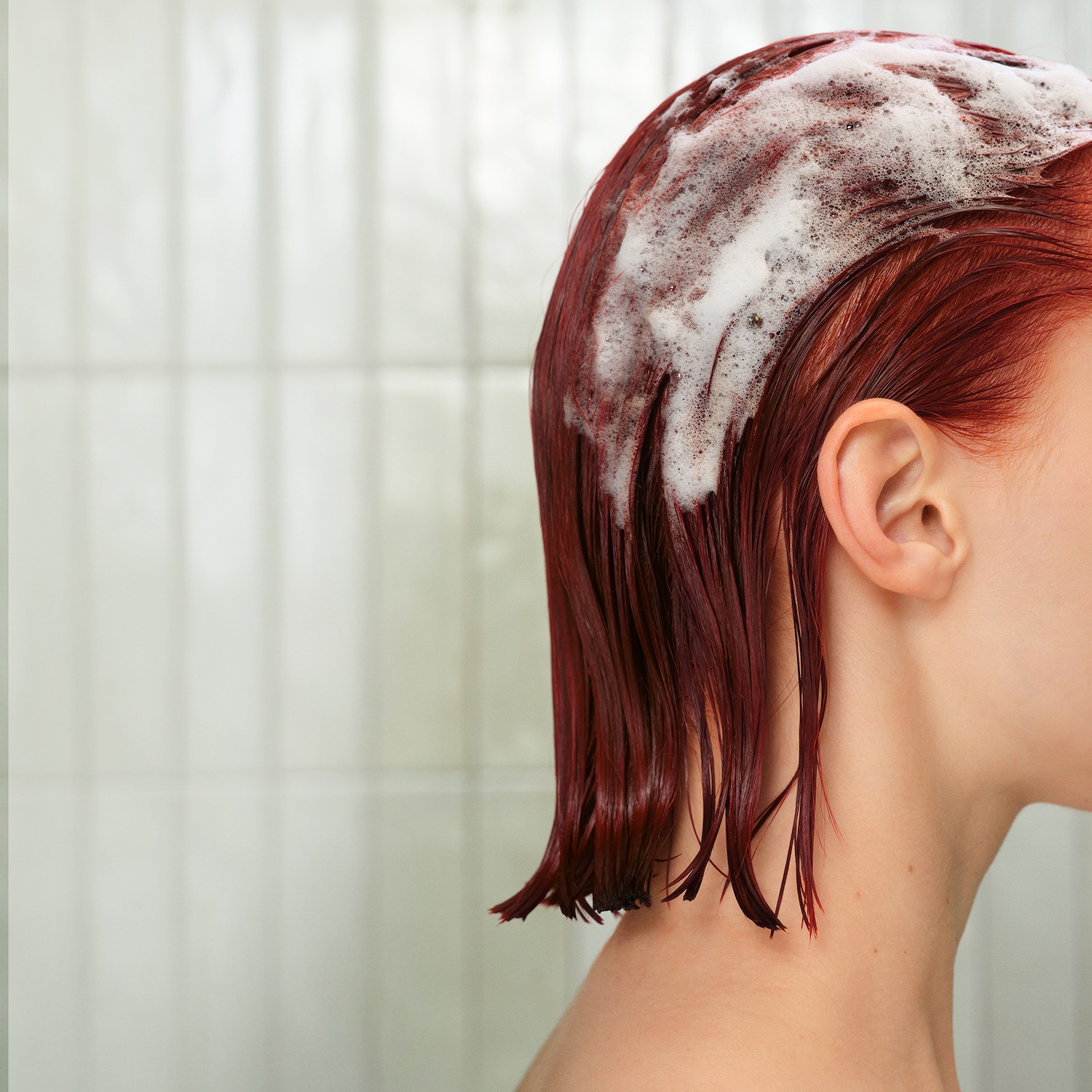 Vista laterale di una donna dai capelli rosso scuro, bagnati e insaponati, che si lava i capelli sotto la doccia.